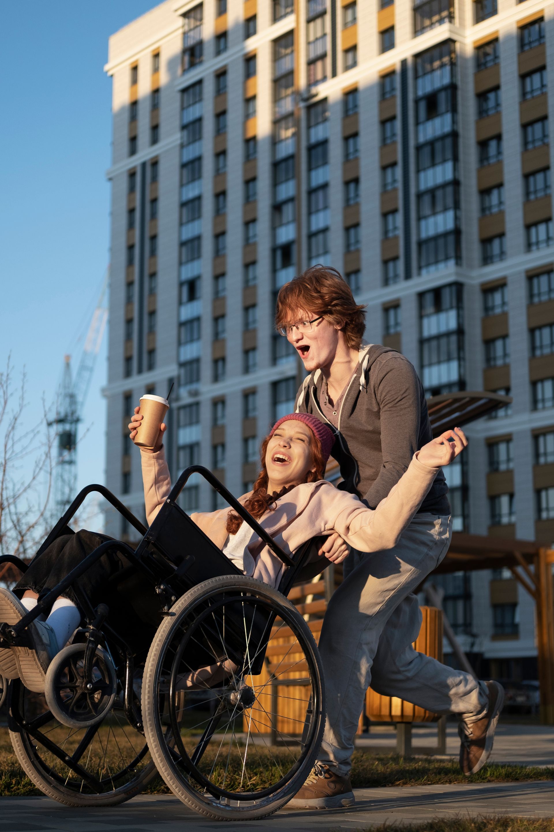 Woman in wheelchair, holding coffee, laughs as a person pushes her outdoors near a building.