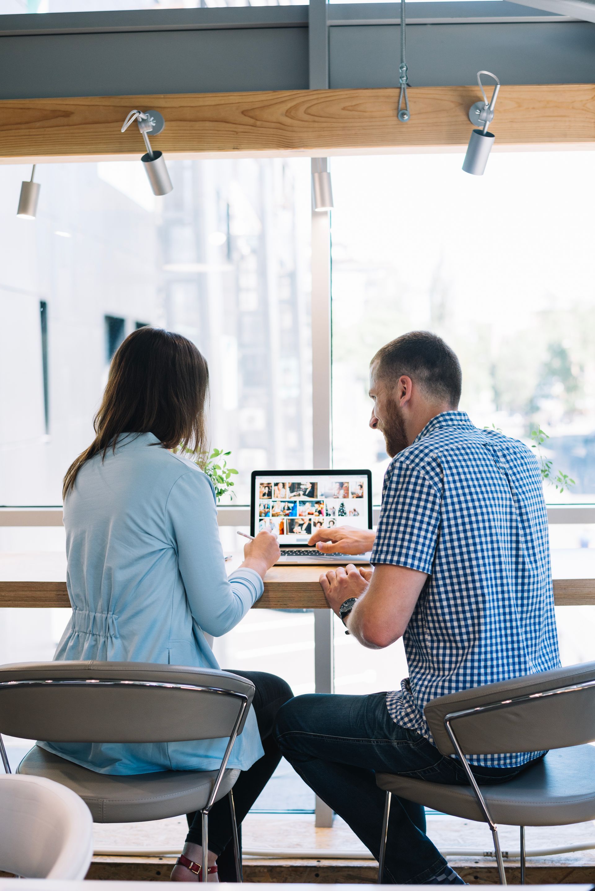 Two people looking at laptop screen in front of a large window, sitting at a desk.