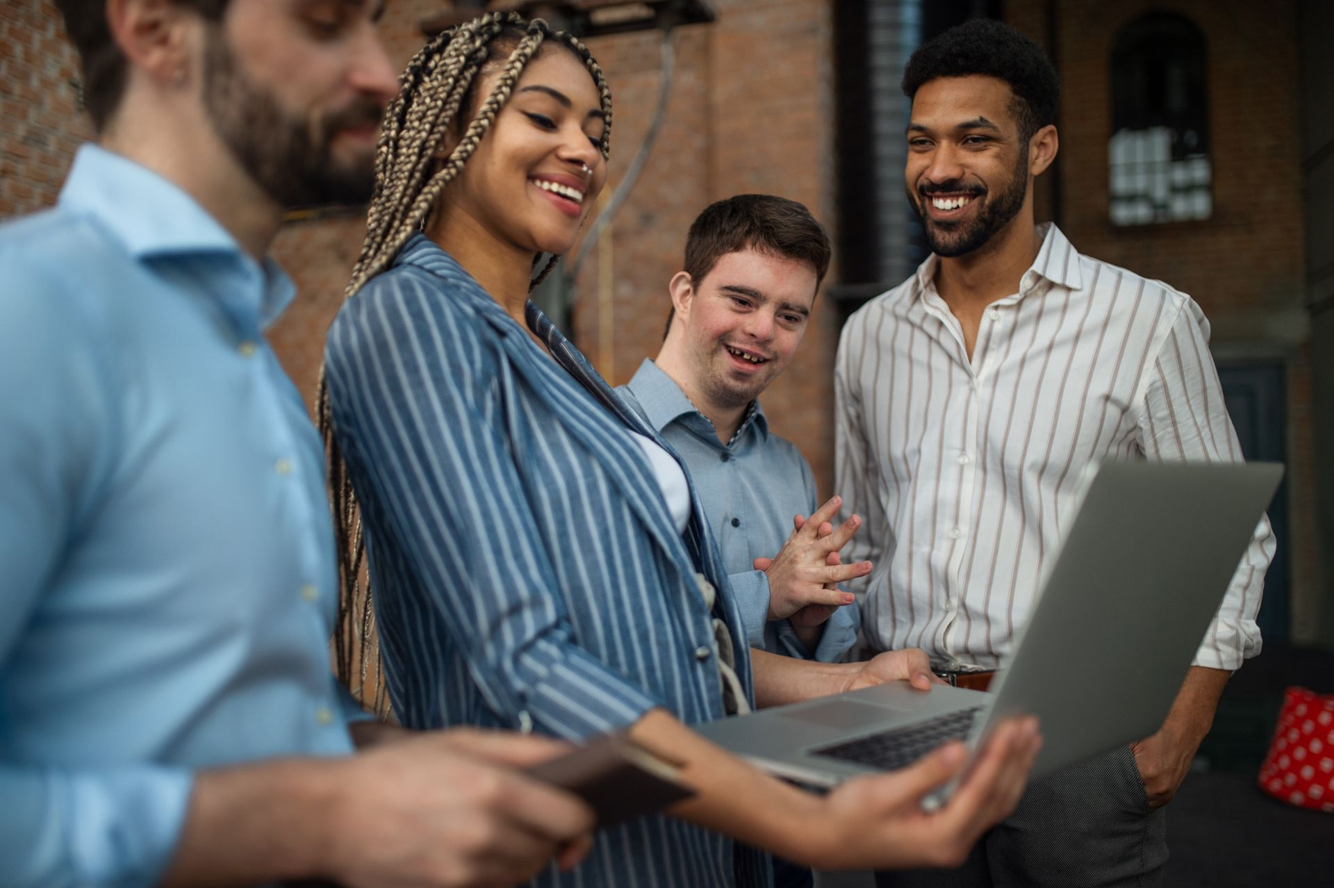 Four people, some smiling, looking at a laptop computer. Outdoors, brick background.