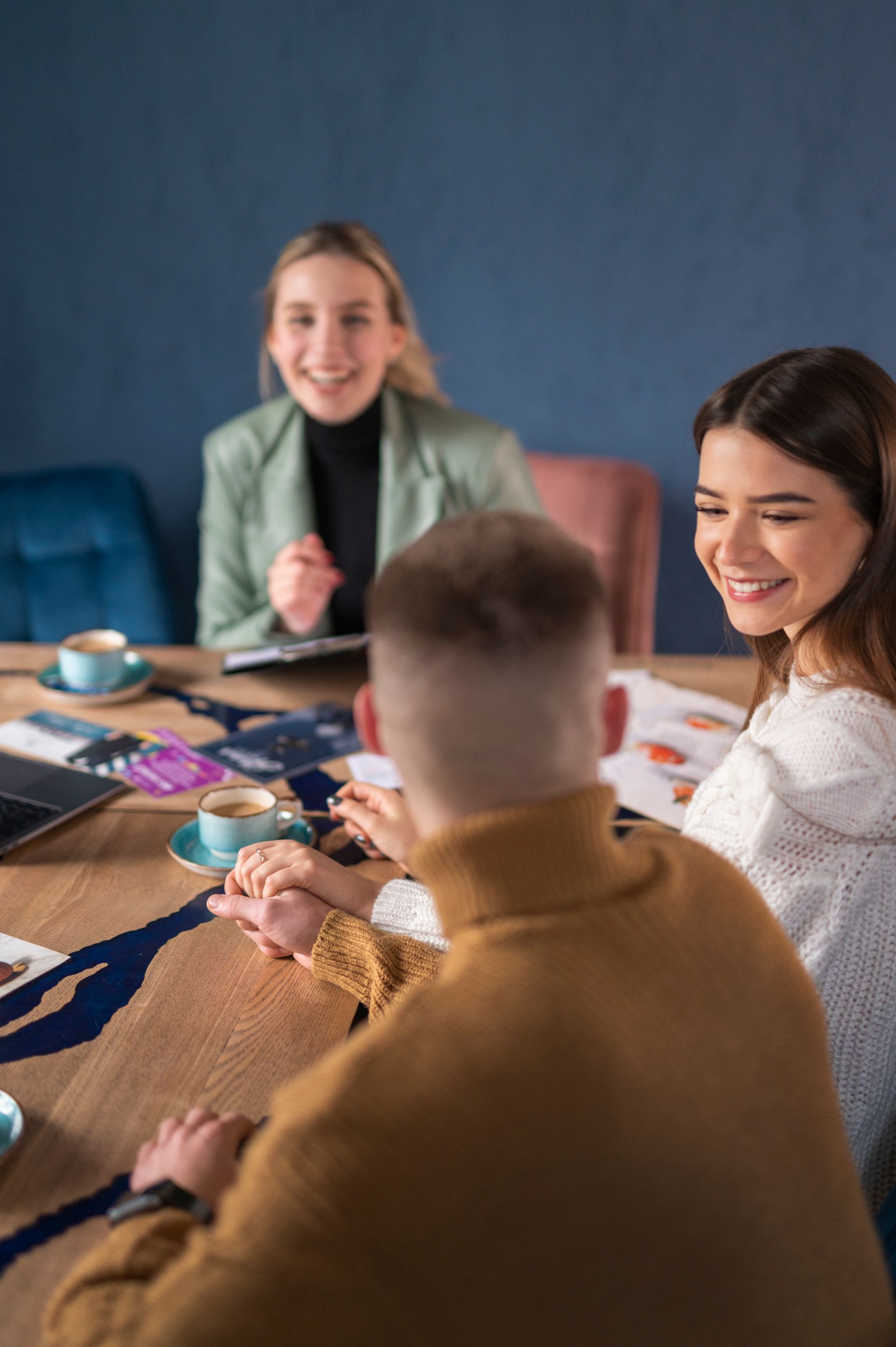 Four people at a table, smiling. One in a brown sweater, another in white, a third in green, and a fourth in the background. Coffee cups are on the table.