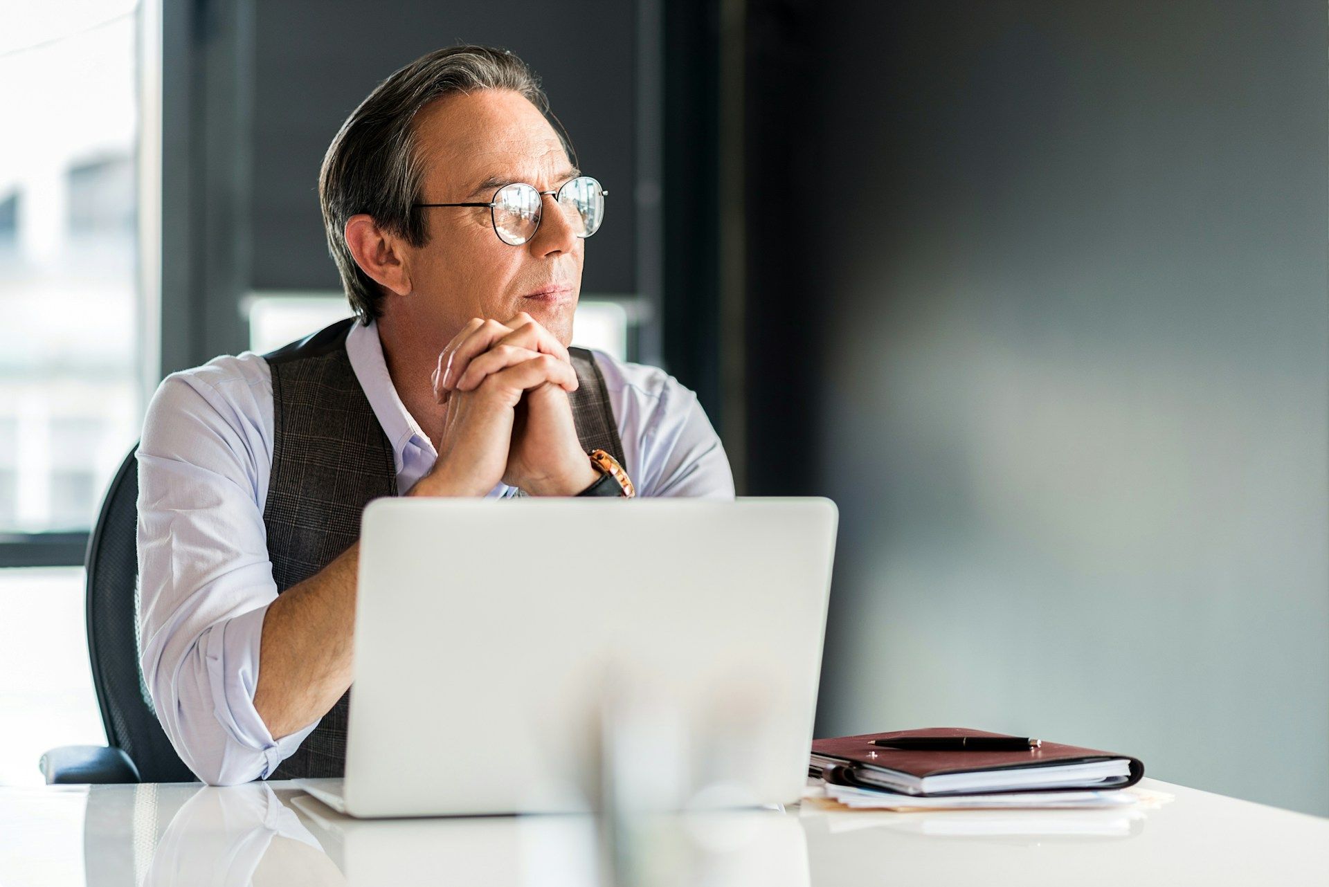 Man wearing glasses at a desk, looking away from a laptop. Hands clasped, thoughtful expression.