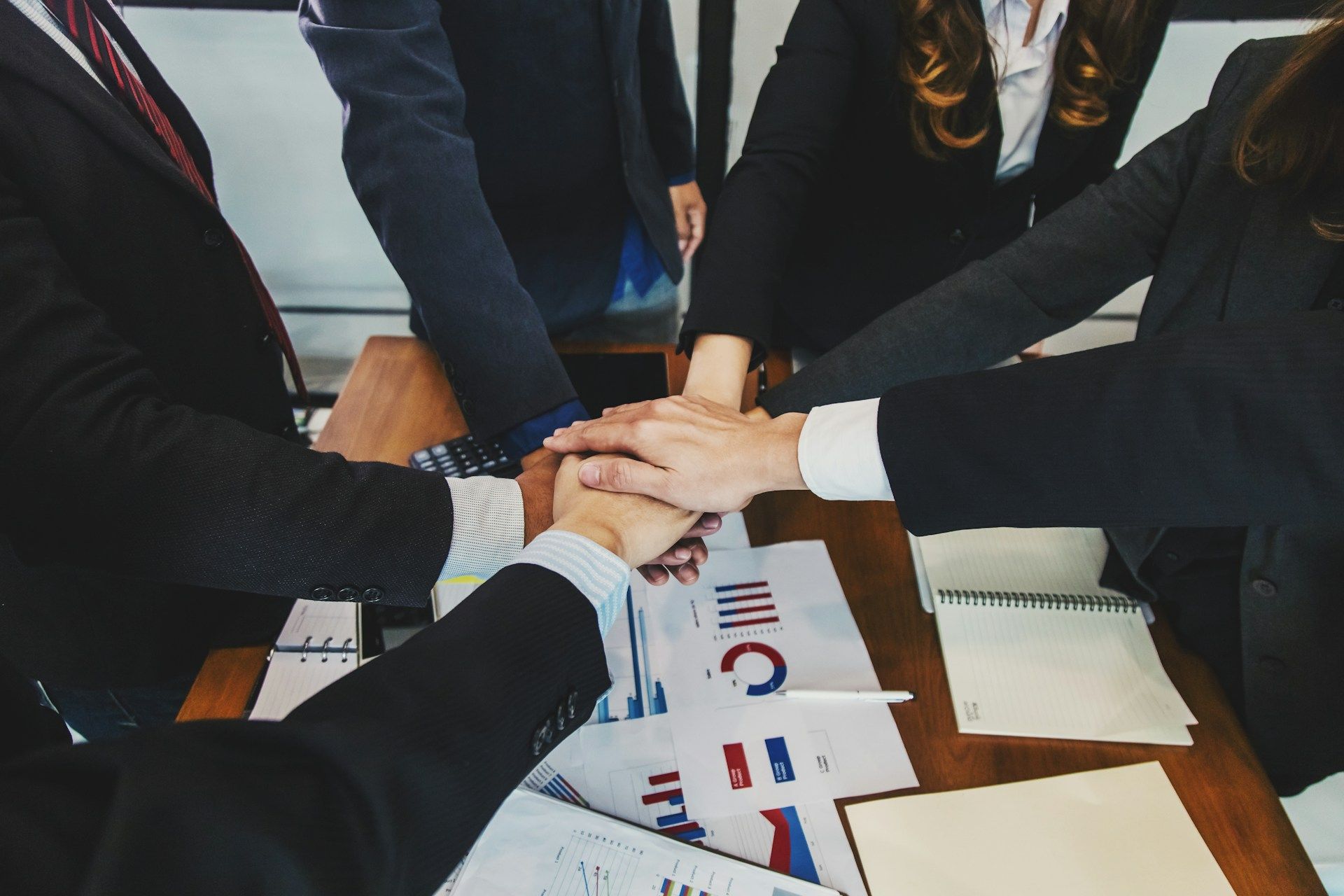 People in business suits huddle, hands together, over a table with papers and charts.