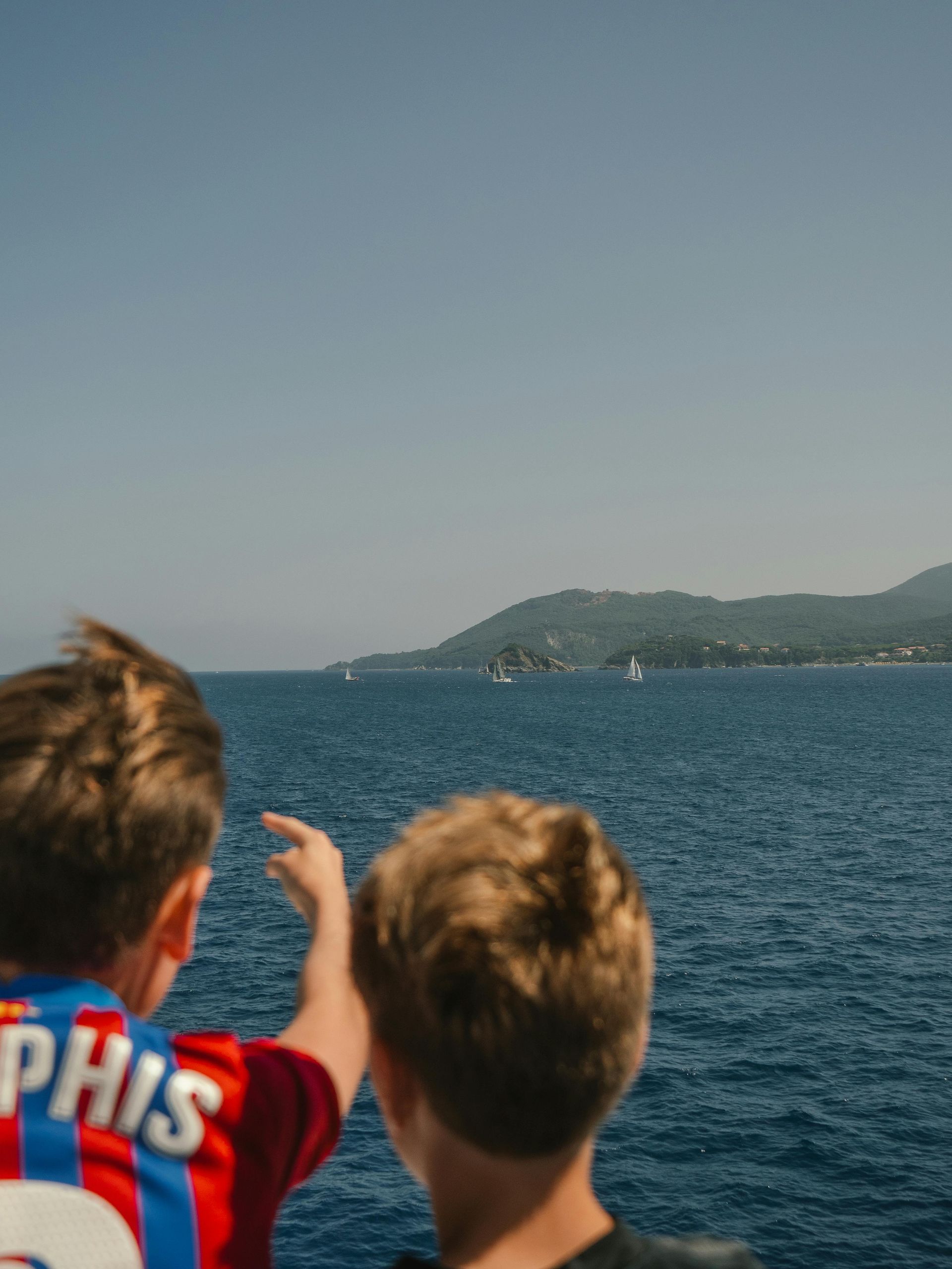 Two boys on a boat look out at the ocean, one pointing towards a distant shore. Blue sky, water, and green land.