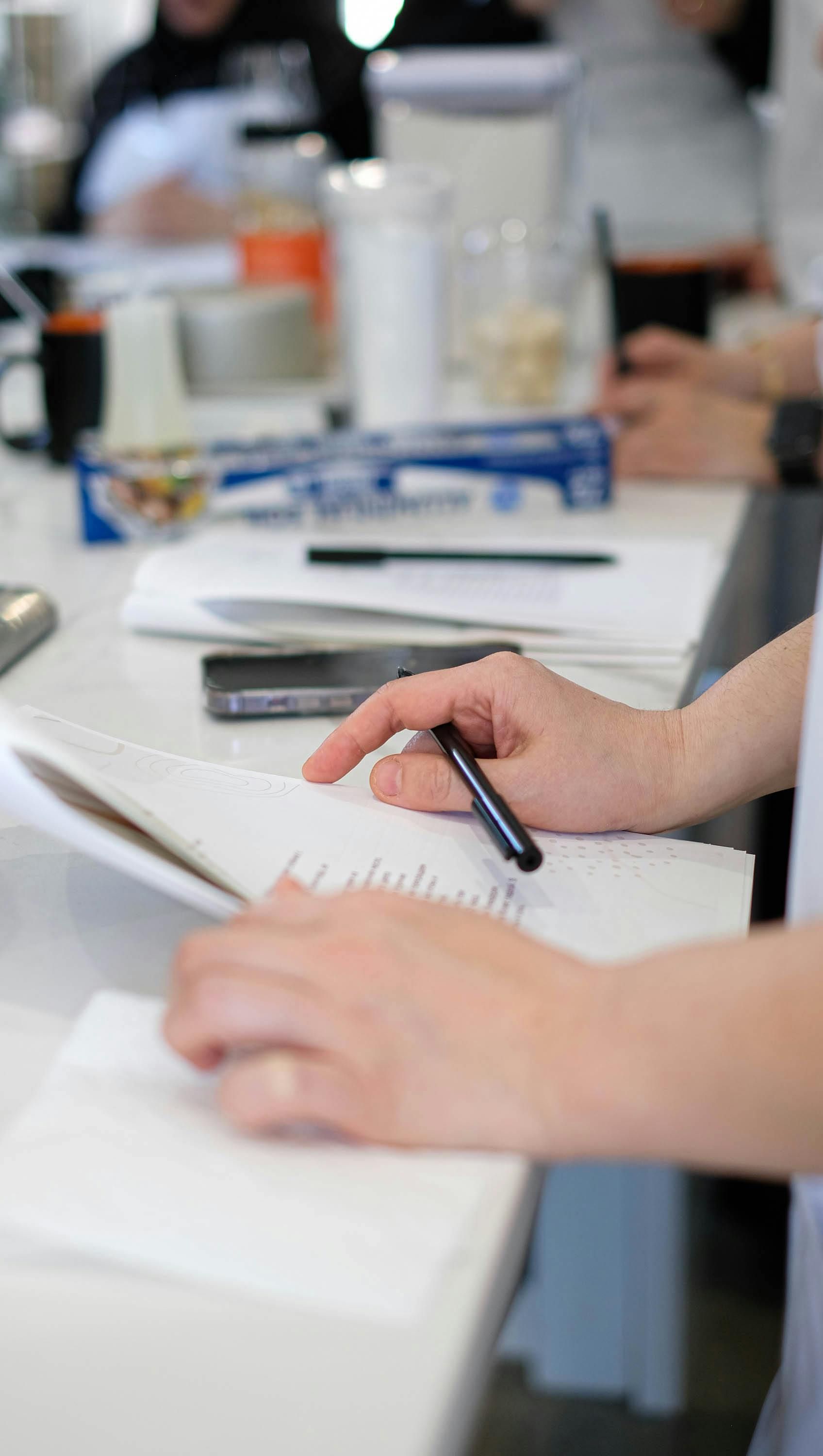 Hands writing in a notebook on a bright kitchen counter with other people and supplies in the background.