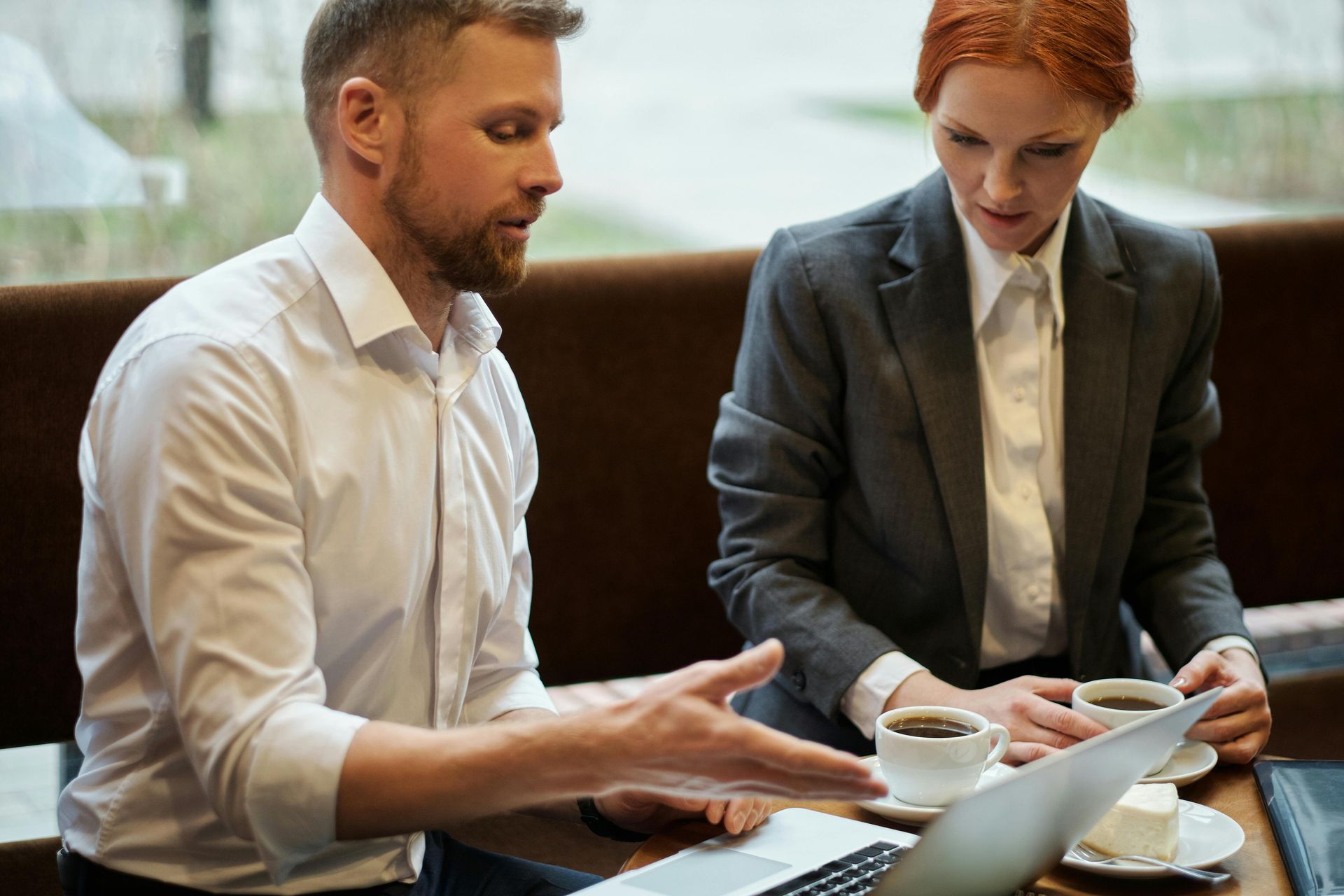 Man and woman at table looking at laptop, both holding coffee cups, discussing work.