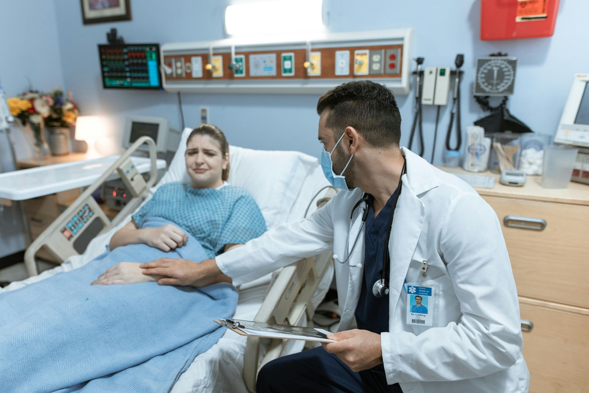 Doctor examining a patient in a hospital bed. The patient looks uncomfortable, doctor wearing a mask. Hospital room.