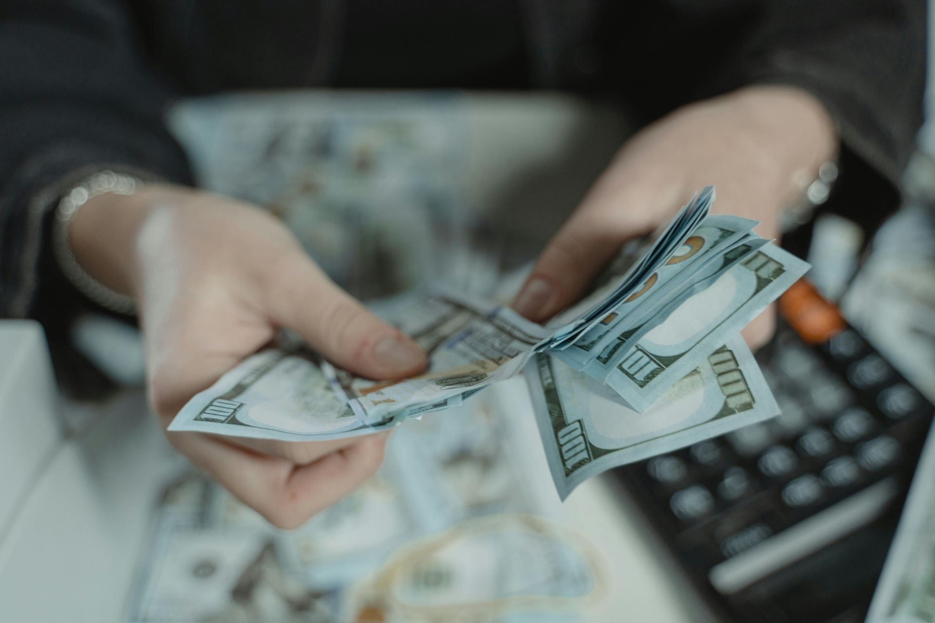 Hands counting a stack of US $100 bills; calculator and more cash are nearby on a white surface.