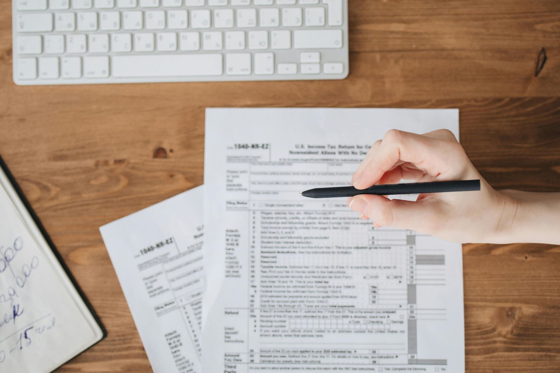 Hand holding a pen over a tax form on a wooden desk with a keyboard and notepad.