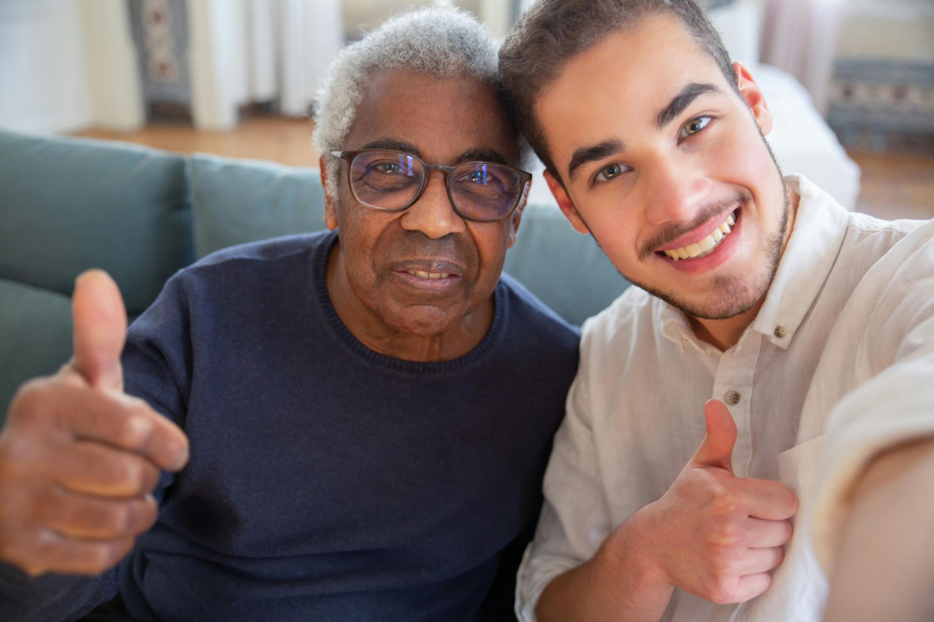 Two people smiling and giving thumbs up, indoors on a sofa.