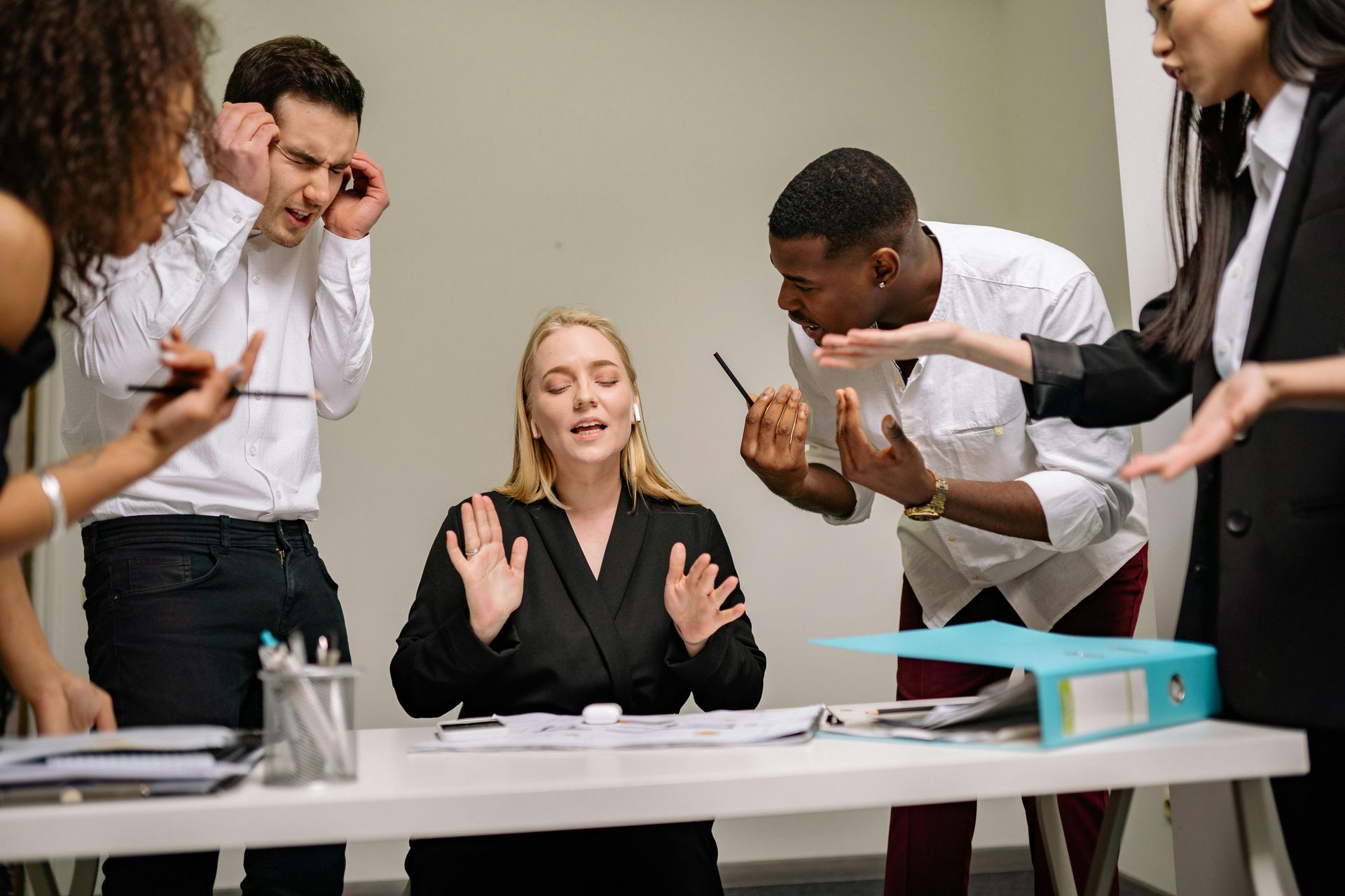 Office coworkers arguing at a desk. Woman in black blazer appears stressed amidst the chaos.