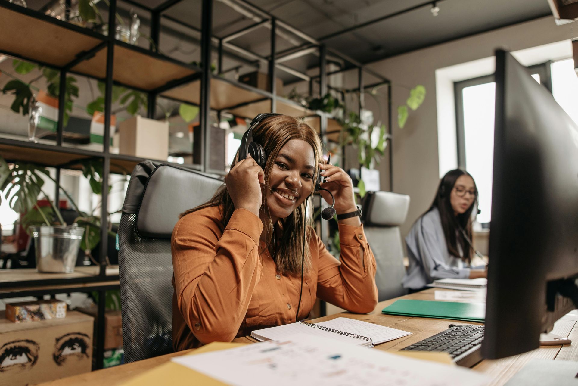 Woman wearing a headset smiles while working at a desk in an office. Another person works in the background.
