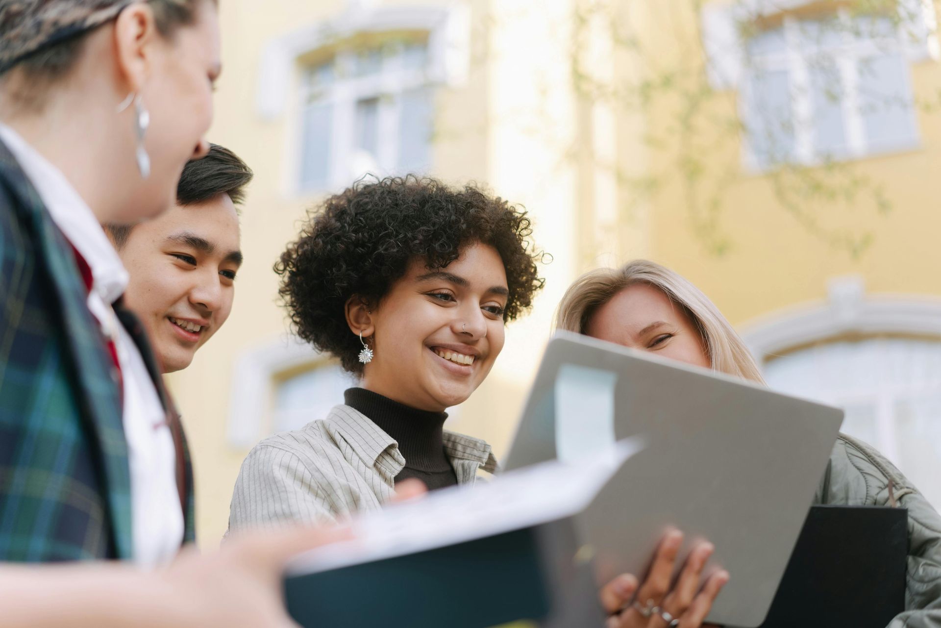 Group of young people smiling, looking at a laptop outdoors near a building.