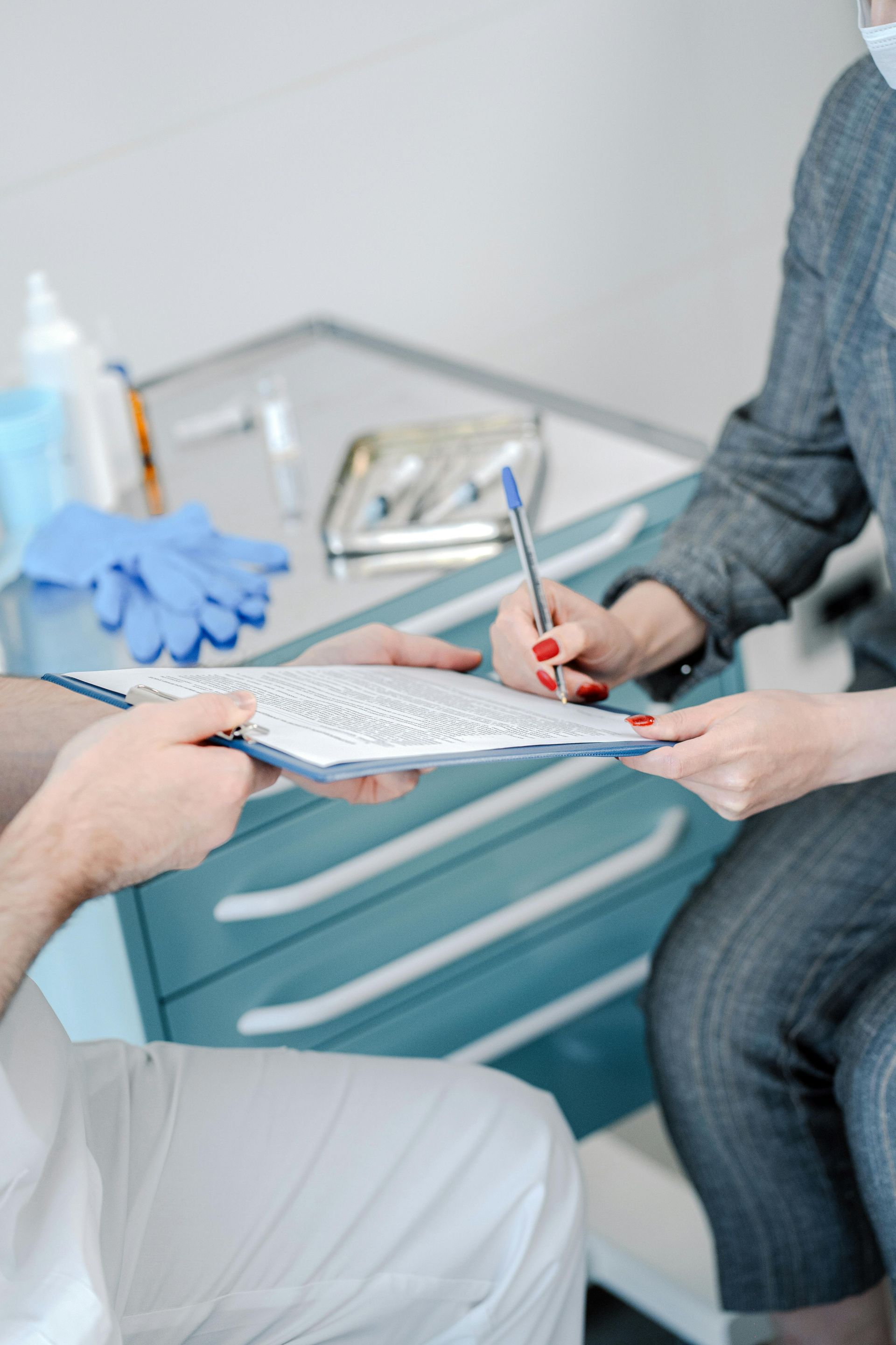 Person signing a document on a clipboard held by another person in a medical setting, surgical gloves and supplies in background.