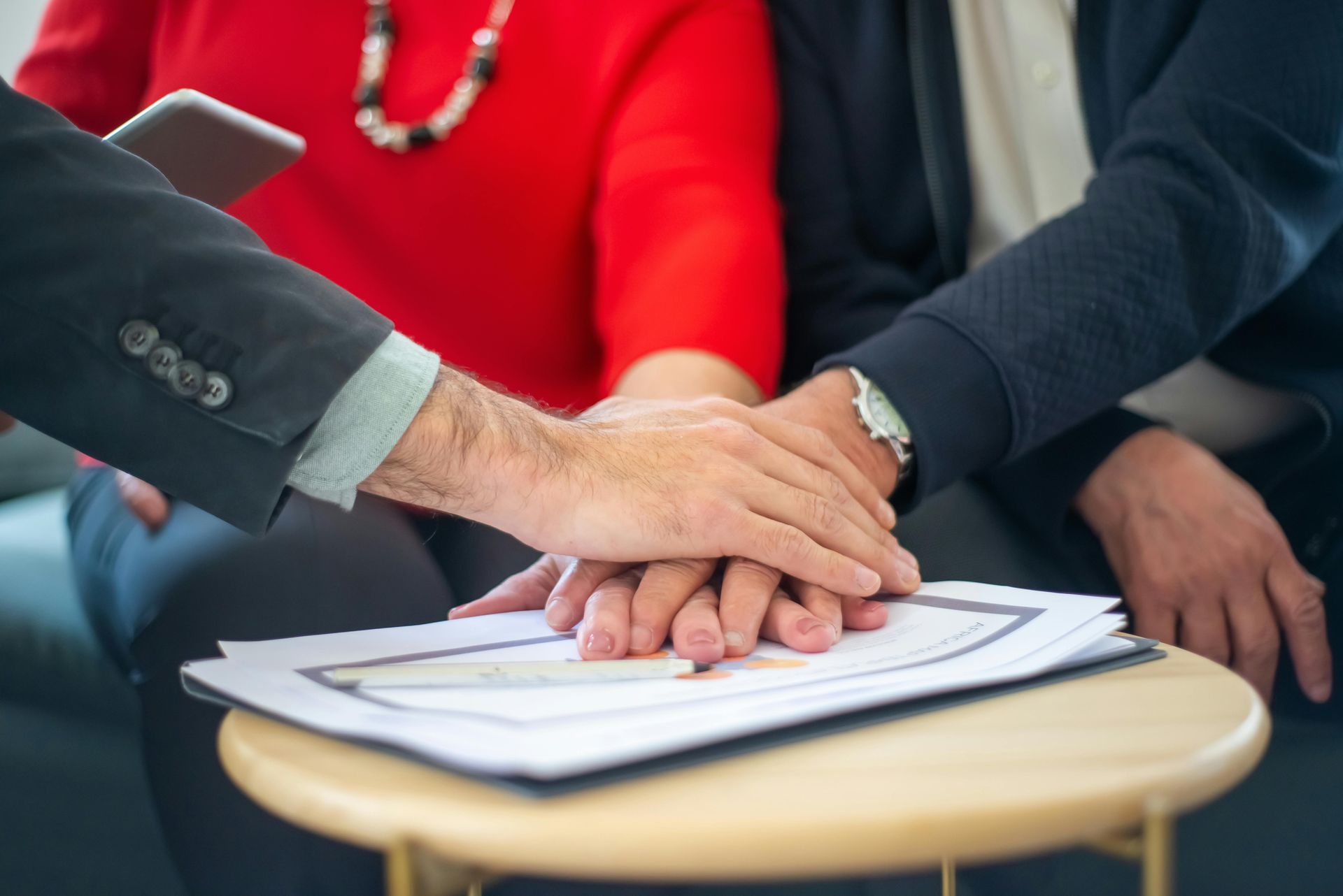 Hands stacked on top of each other over documents. A person in a red shirt sits next to another person with a navy blue shirt.