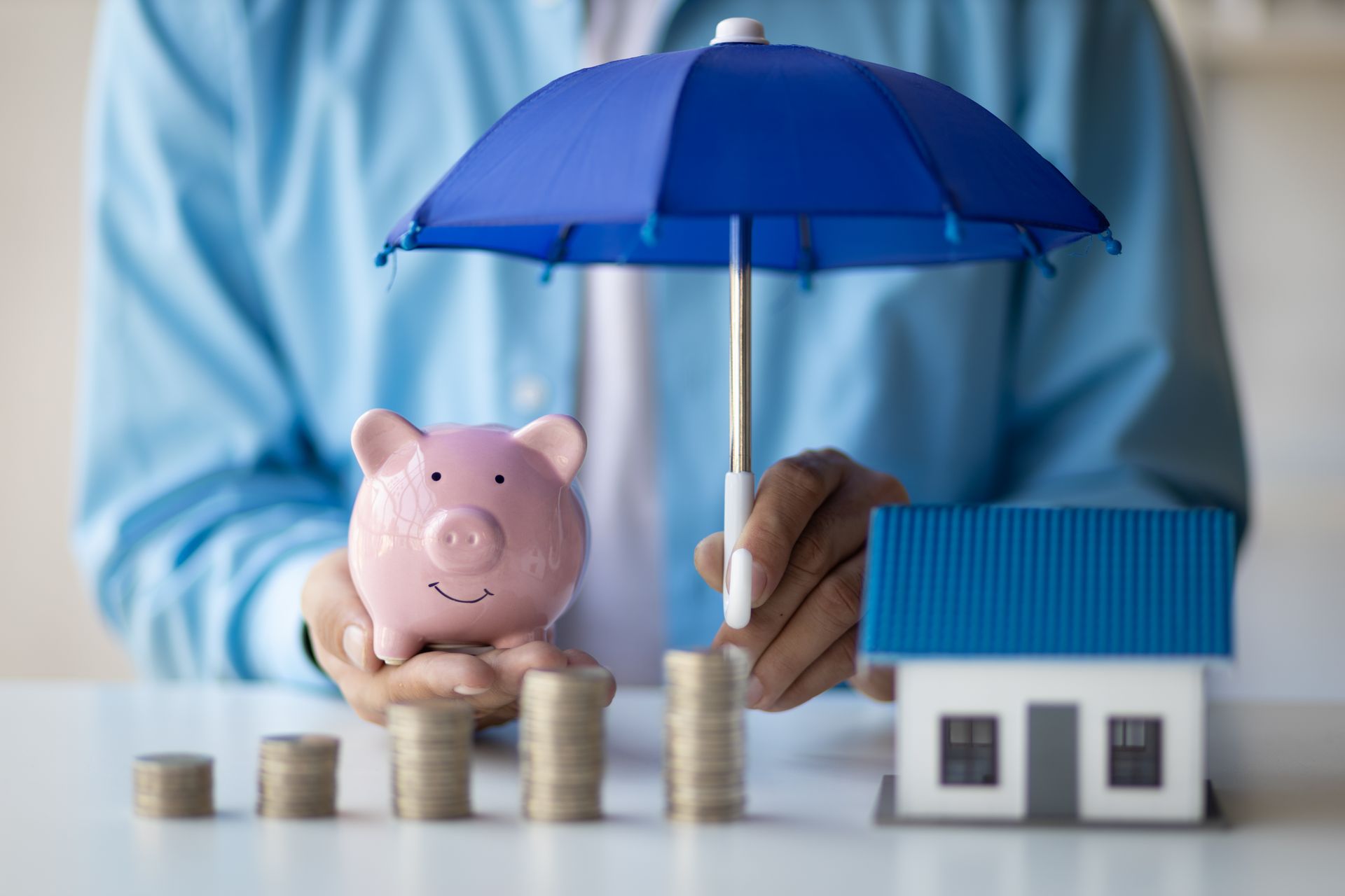 Person holding a blue umbrella over a piggy bank, stacks of coins, and a house model, symbolizing financial protection.