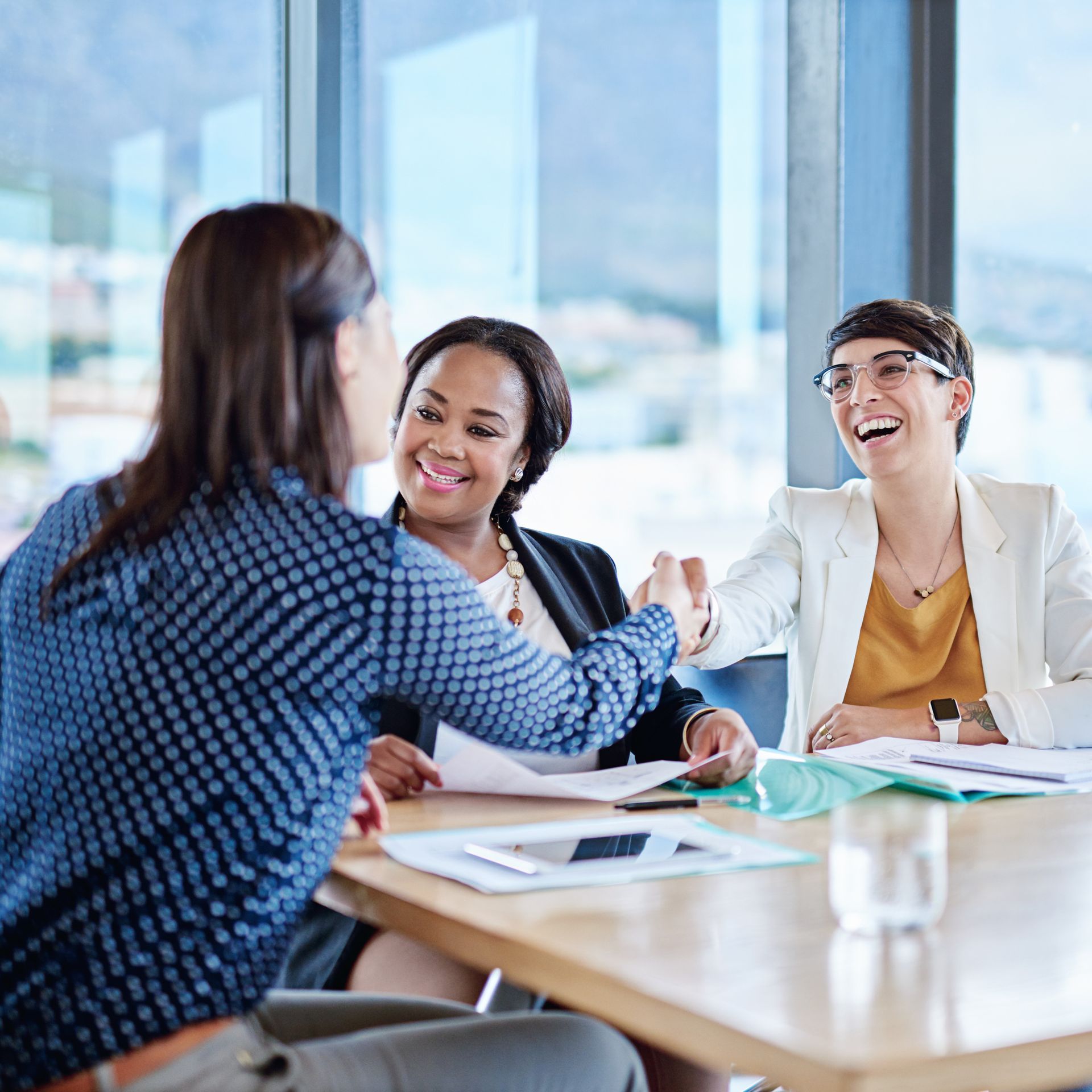 Three women at a table; one is shaking hands, the others smile. Papers and tablet are on the table in a bright office.