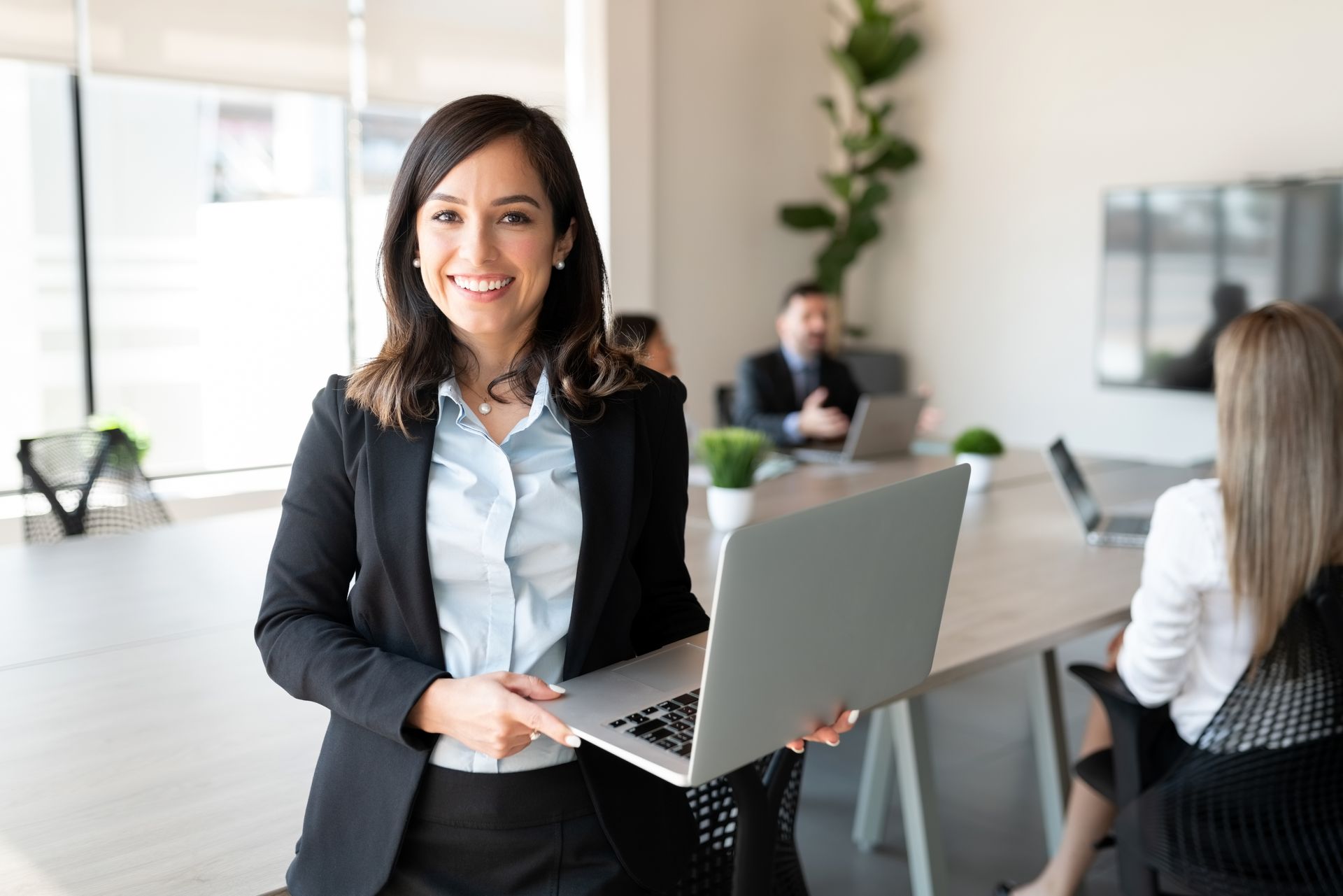 Woman in business attire smiles, holding a laptop in an office setting.