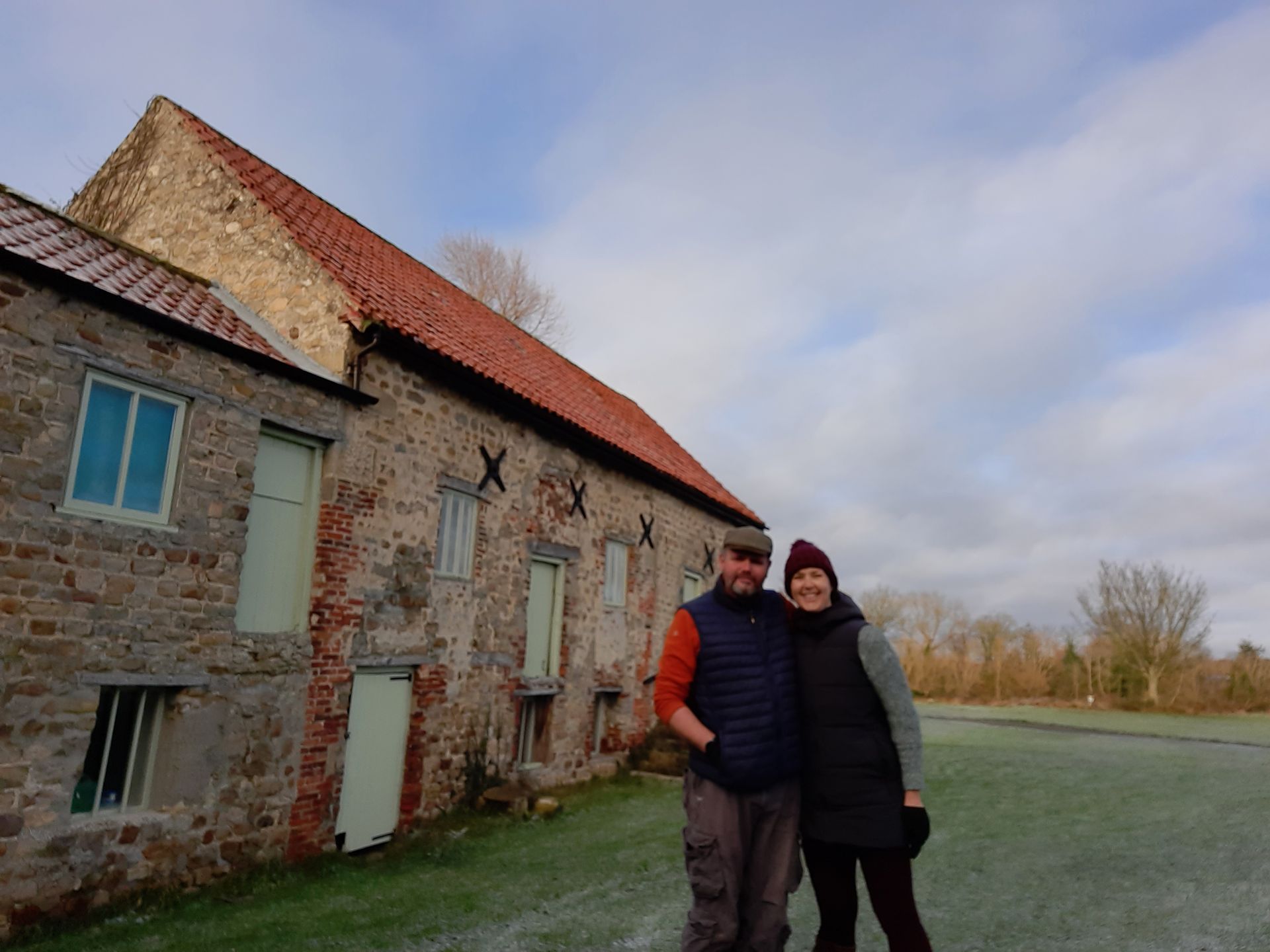 Couple standing in front of an old stone building with a red tiled roof, on a cloudy day.