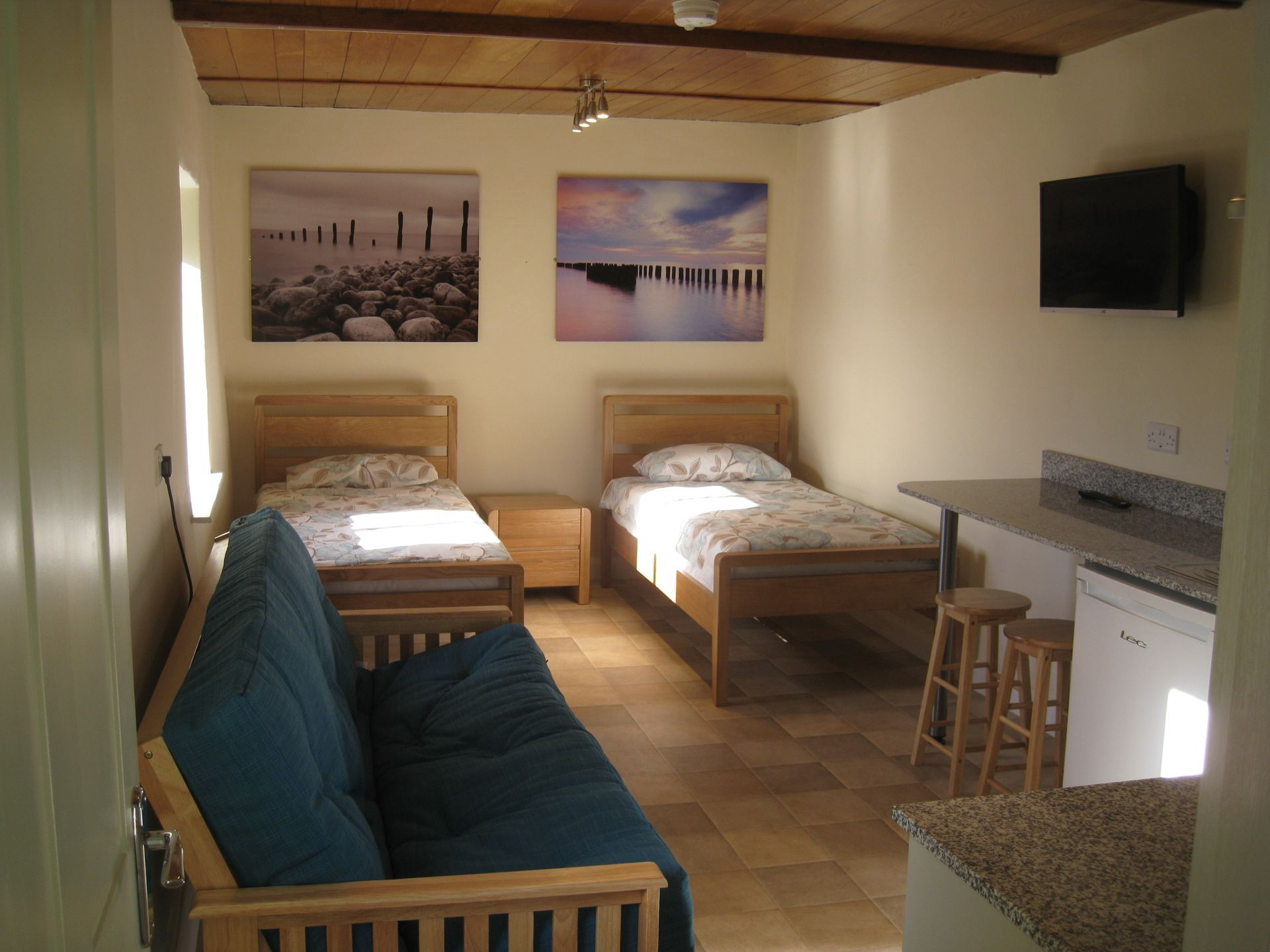 Small room with two beds, futon, kitchenette, and artwork. Brown wood furnishings and pale walls.