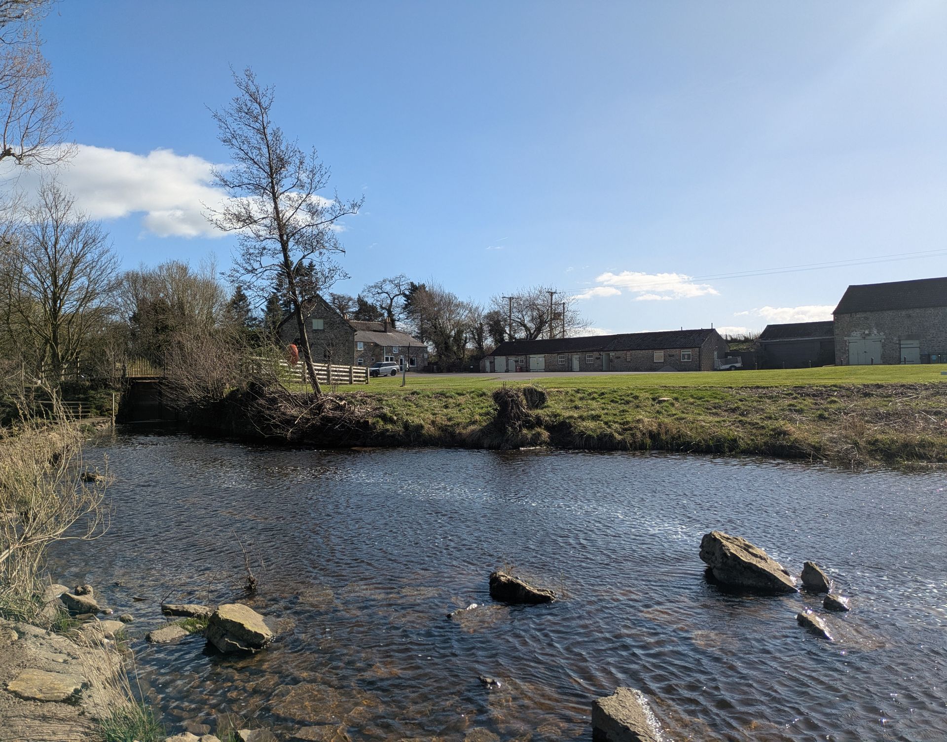 A stream flows towards a grassy field and buildings under a bright blue sky.