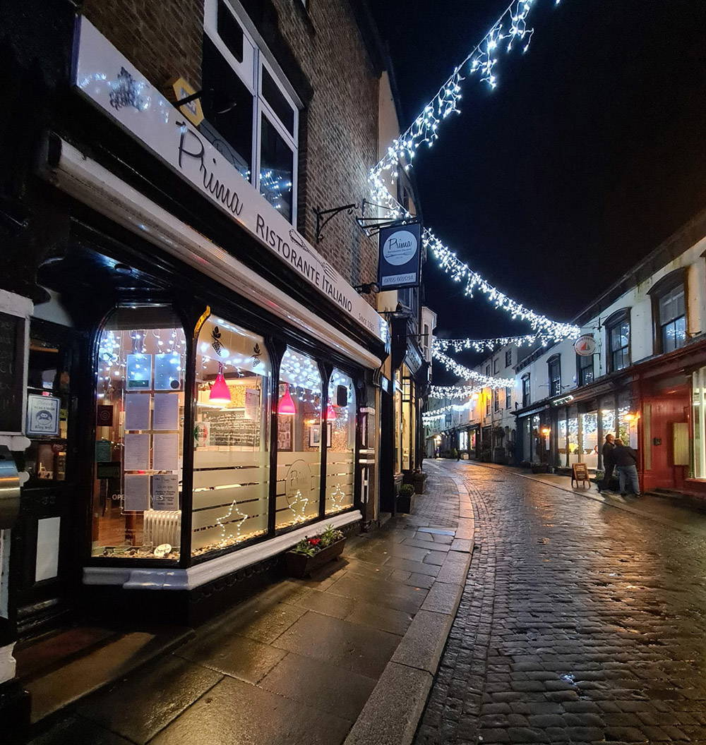 Nighttime view of a cobblestone street lined with shops, lit with string lights.