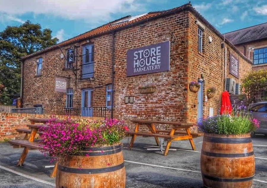 Store House restaurant exterior with brick walls, picnic tables, and barrels with purple flowers.