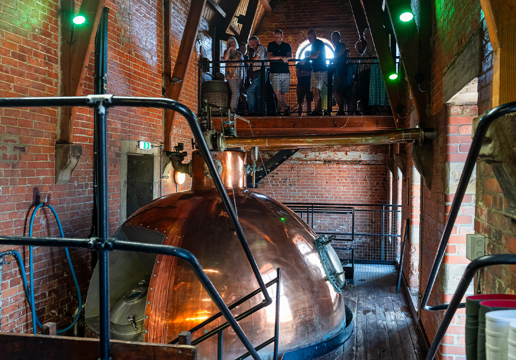 Large copper brewing vat inside a brick building, with people observing from a wooden balcony above.