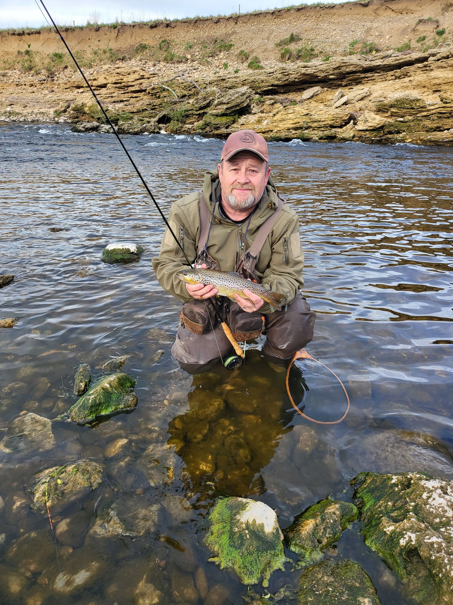 Man in waders kneels in river, holding a fish. Fishing rod overhead. Rocky riverbed, tan cliffs in background.