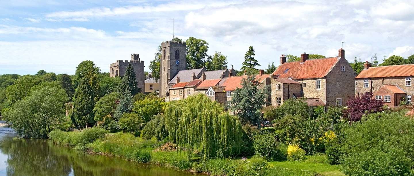 A riverside town with stone buildings, red-tiled roofs, and a tall church tower, surrounded by lush green trees under a blue sky.