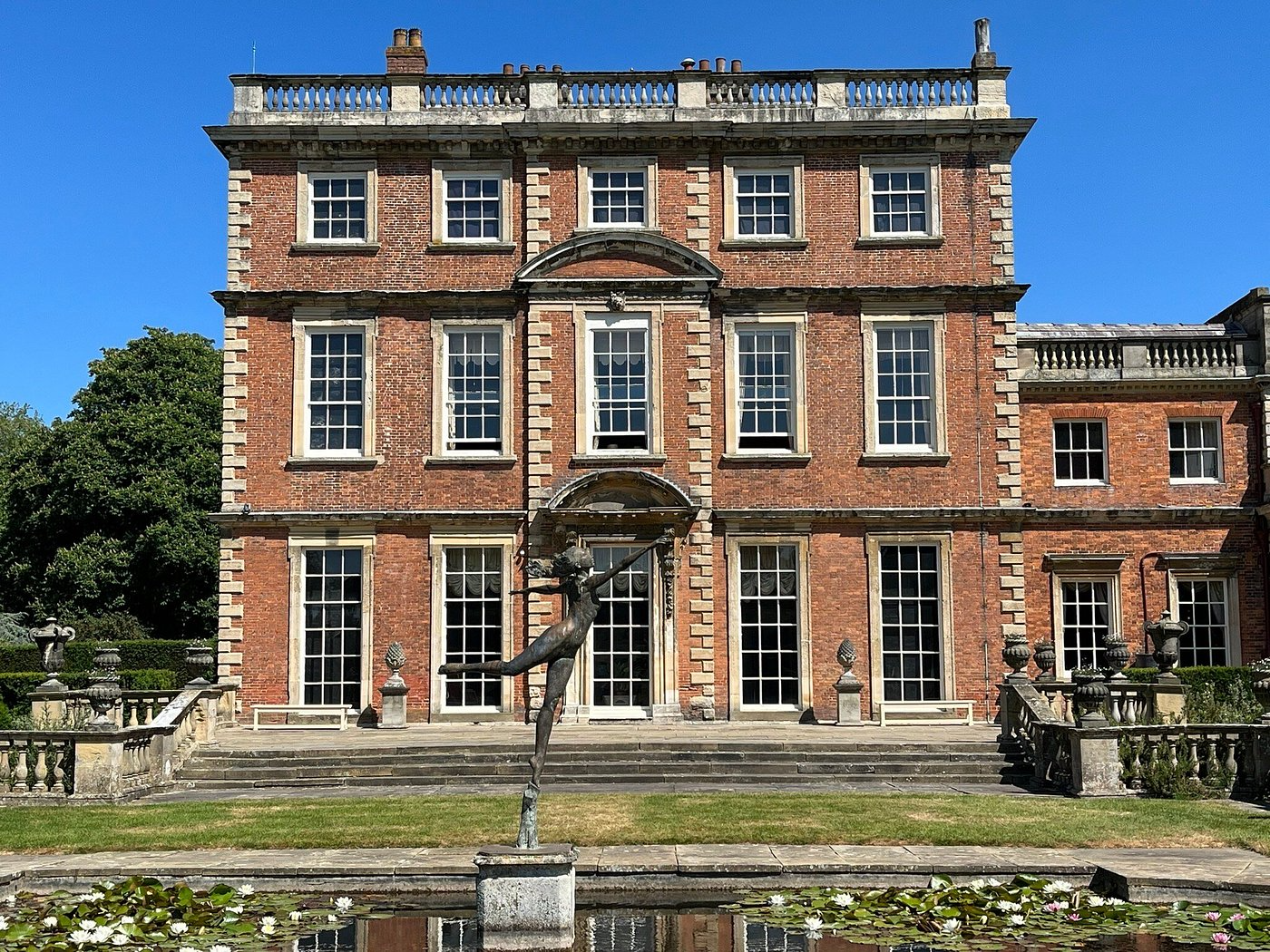 Brick manor house with white trim, statue in foreground, reflecting pool.