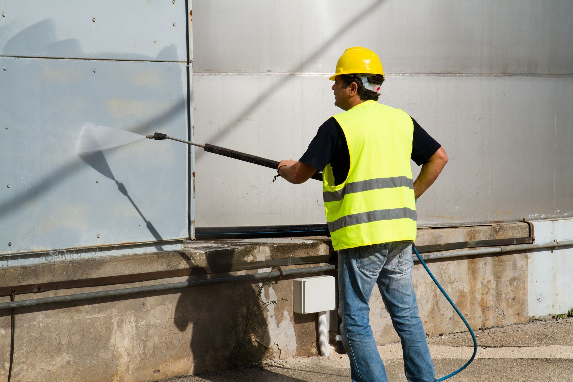 A man in a yellow vest and hard hat is cleaning a wall with a high pressure washer