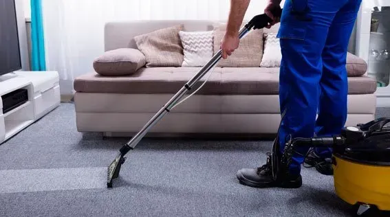 Un trabajador aspira una alfombra gris en una sala de estar con un sofá beige y una aspiradora amarilla.