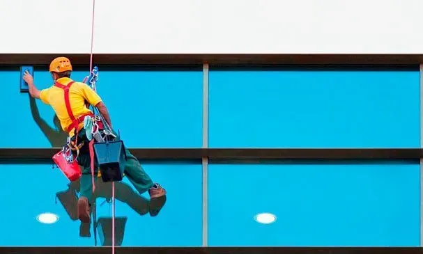 Limpiador de ventanas suspendido por cuerdas junto a la fachada de un edificio de cristal azul.