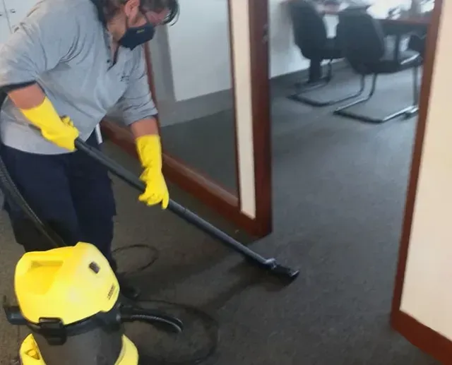 Una persona con guantes amarillos aspira el pasillo alfombrado de una oficina con una aspiradora de bote amarilla.
