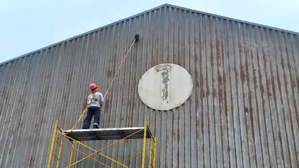 Un trabajador, subido a un andamio, pinta con aerosol un gran letrero ovalado blanco sobre la pared de un edificio de chapa ondulada.
