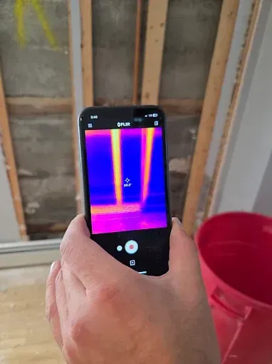 Person holding a phone displaying a thermal image of a ceiling; revealing heat differences. A red bucket is in the foreground.