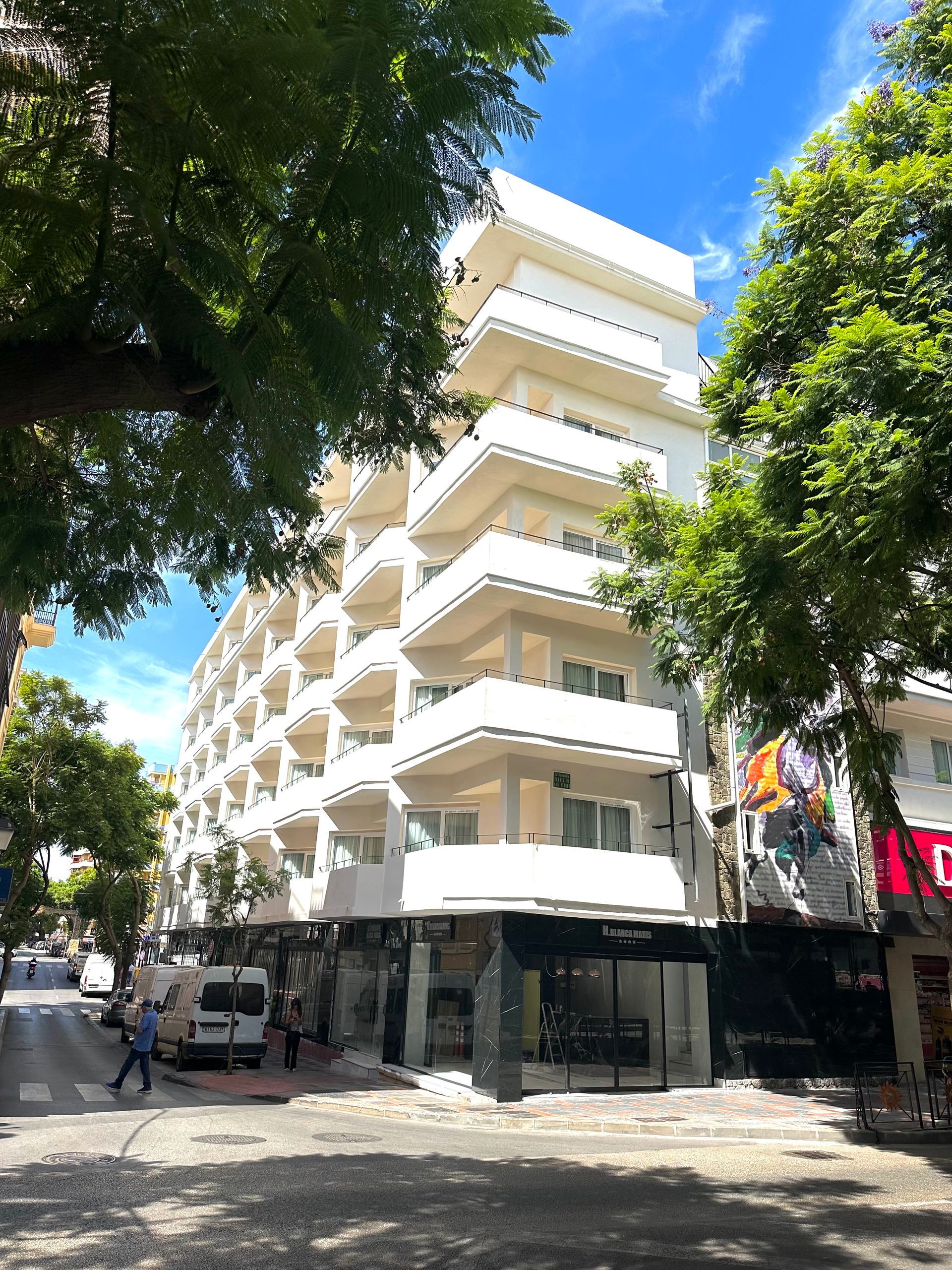 Edificio blanco de varias plantas con balcones, acera, árboles y peatones en la calle.