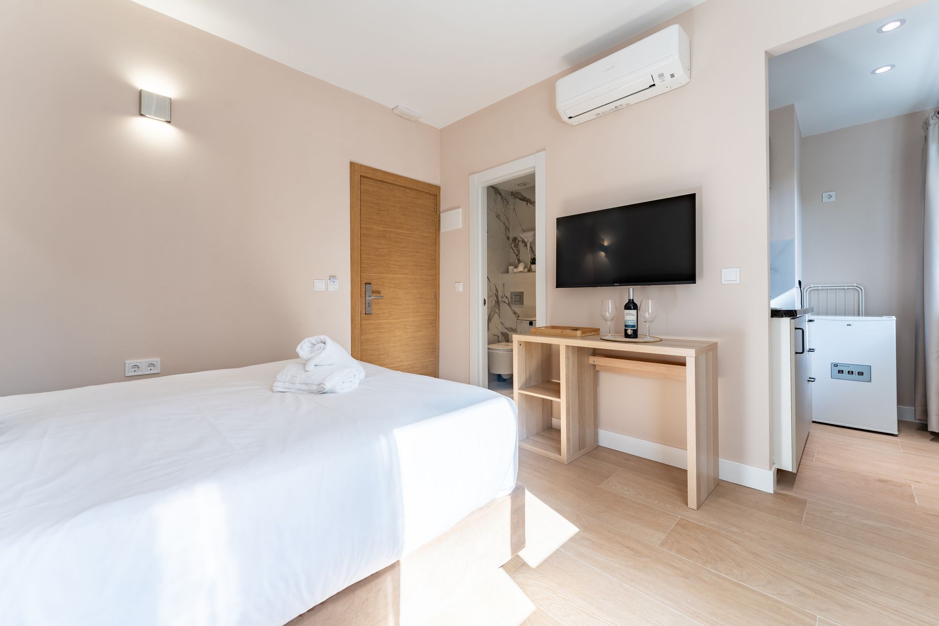 Dormitorio con cama blanca, TV, escritorio de madera y puerta abierta a un pequeño baño.