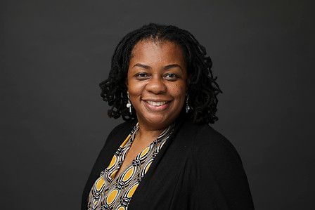Woman with curly dark hair smiles, wearing patterned blouse and black cardigan against a gray backdrop.