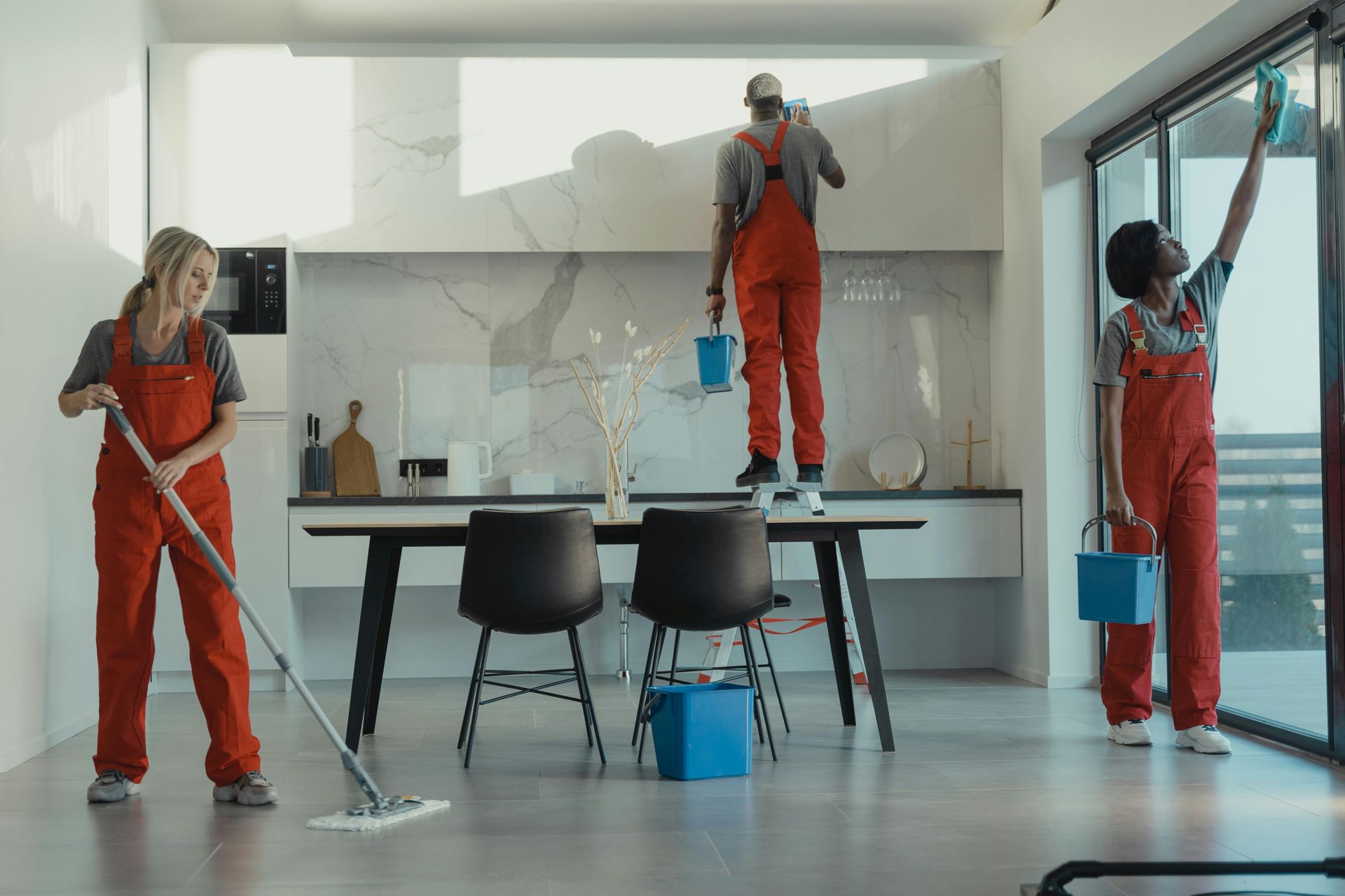 Three people in red jumpsuits cleaning a modern kitchen; one mopping, one painting, and one wiping a window.