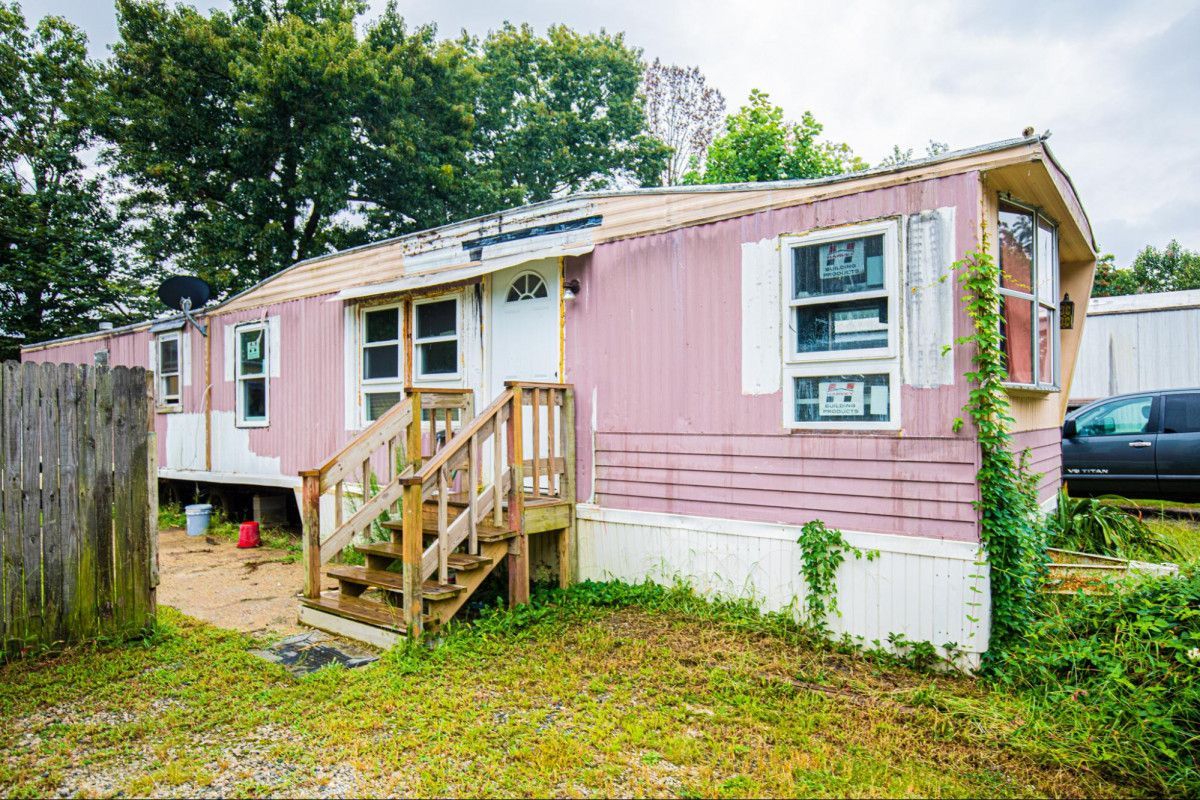 Pink mobile home with peeling siding, wooden steps, and overgrown grass.