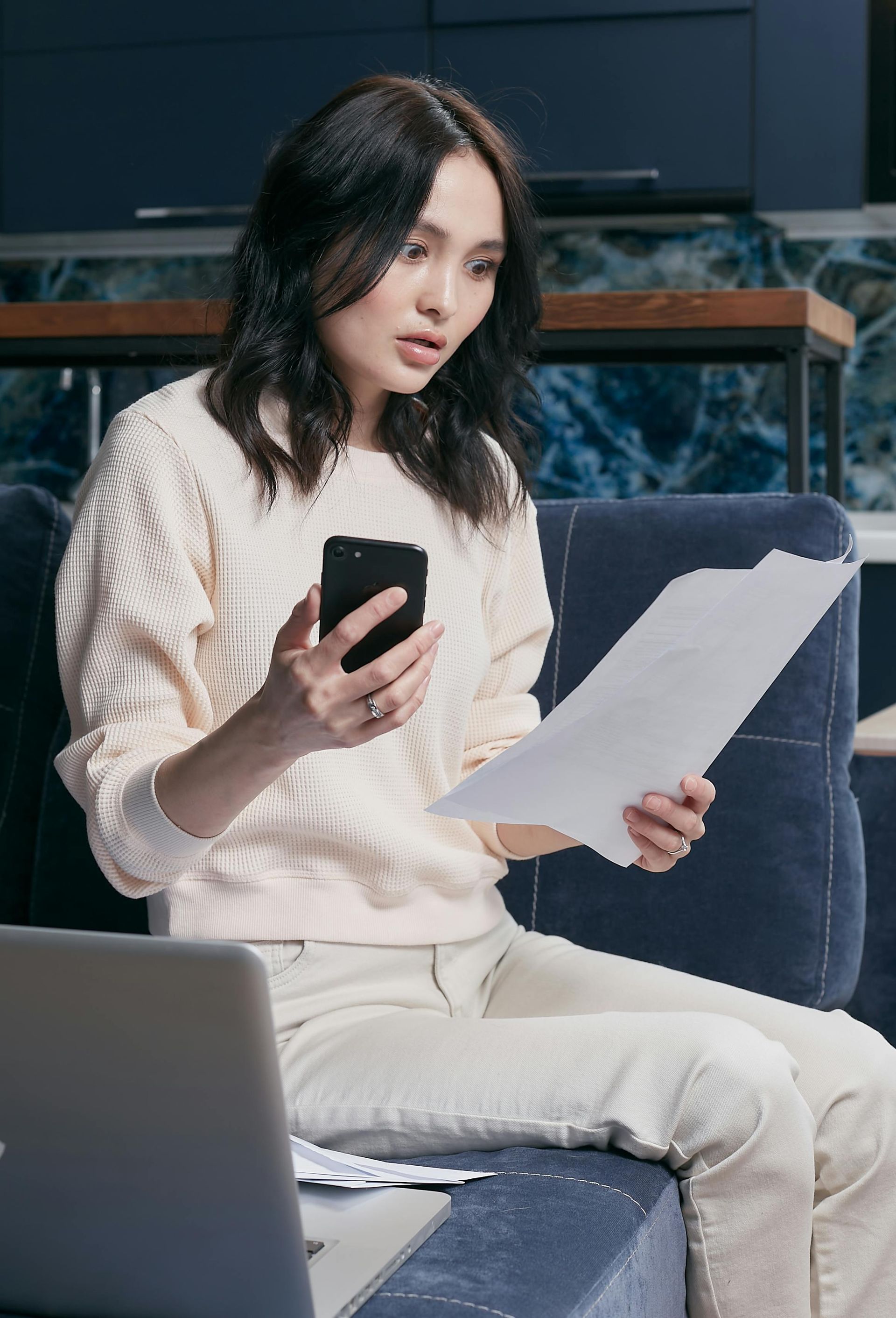 Woman holding a phone and paperwork, looking concerned, sitting on a sofa with a laptop.