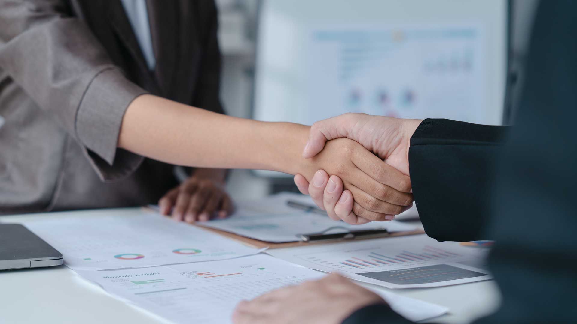 Two people in business attire shaking hands over a desk with documents.