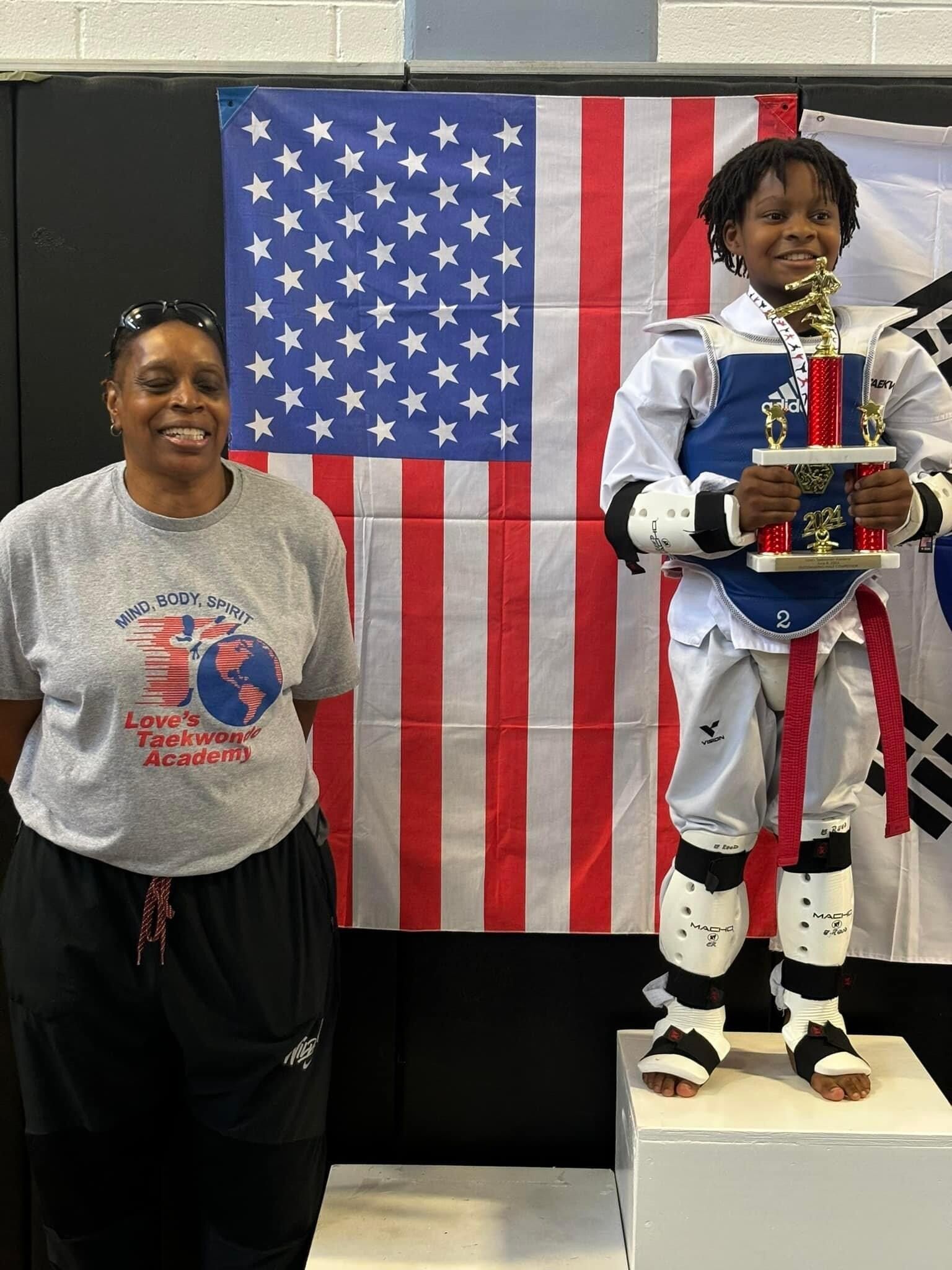 A woman is standing next to a young boy holding a trophy in front of an american flag.