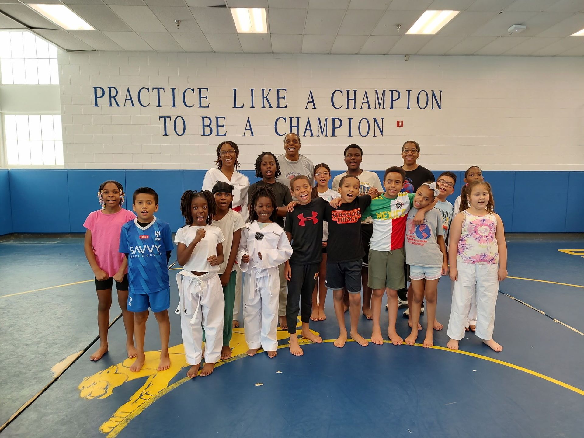 Group of children and adults posing on a wrestling mat in a gym with the text, “Practice like a champion to be a champion.”