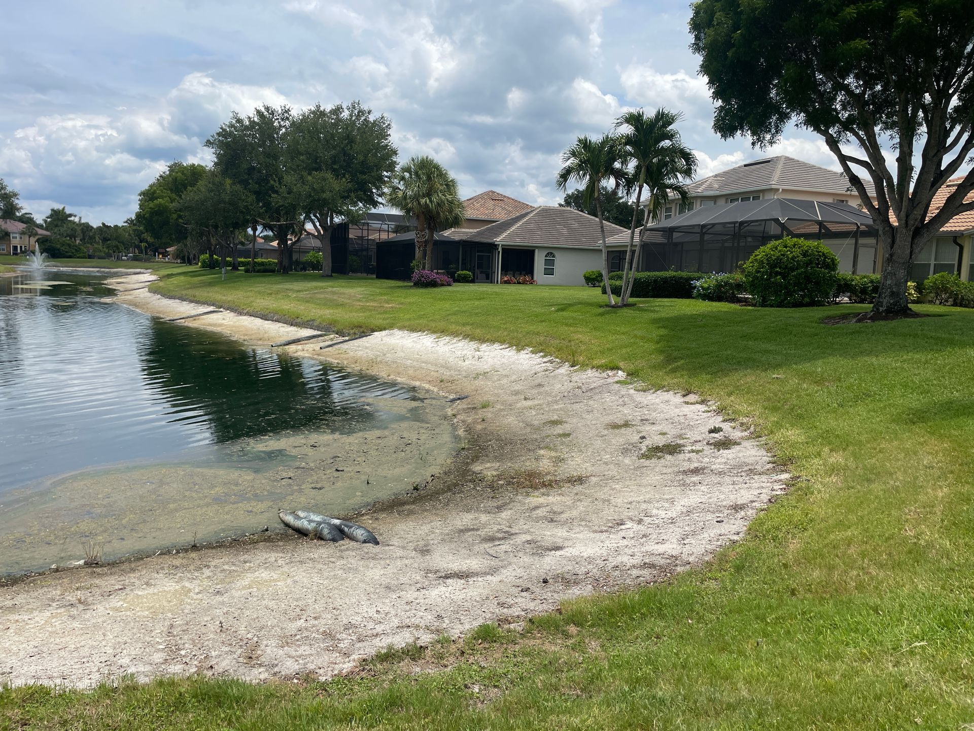 Calm residential canal with grassy bank, sandy shore, and houses under cloudy sky