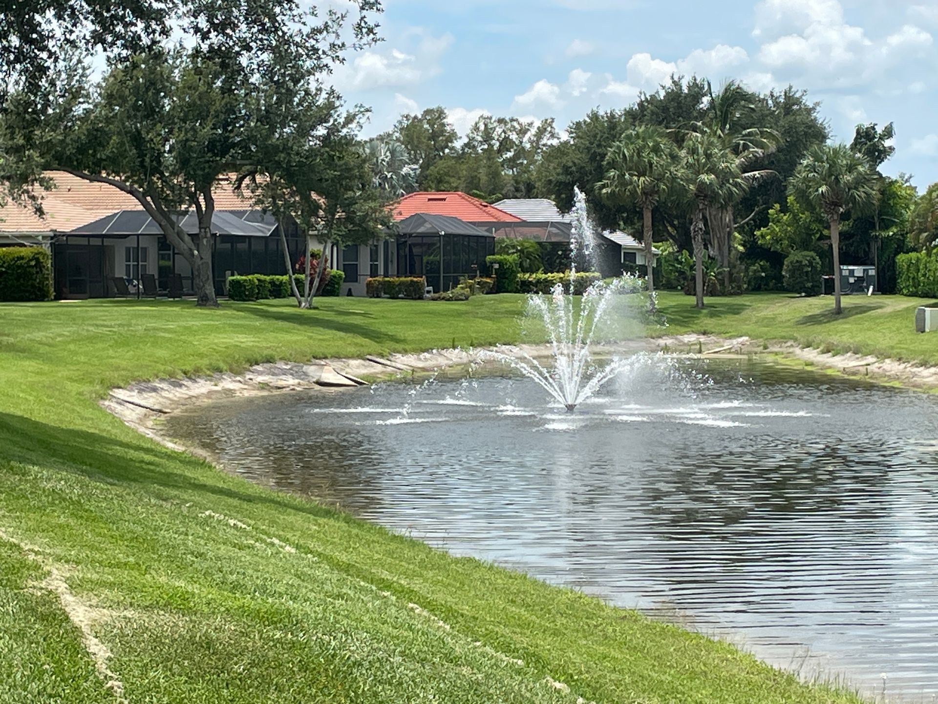 Suburban pond with a fountain, green lawns, trees, and houses under a partly cloudy sky