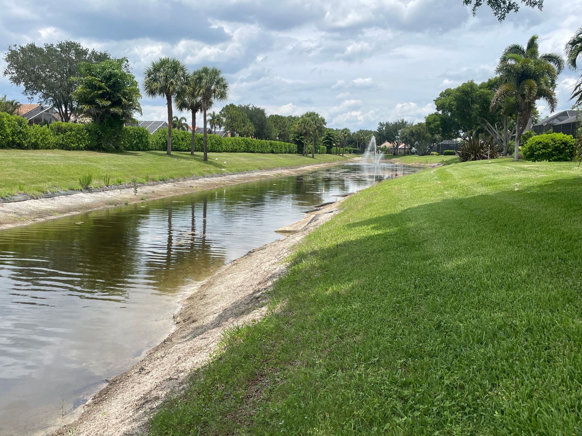 Canal beside a green grassy embankment under a cloudy sky, lined with trees and shrubs