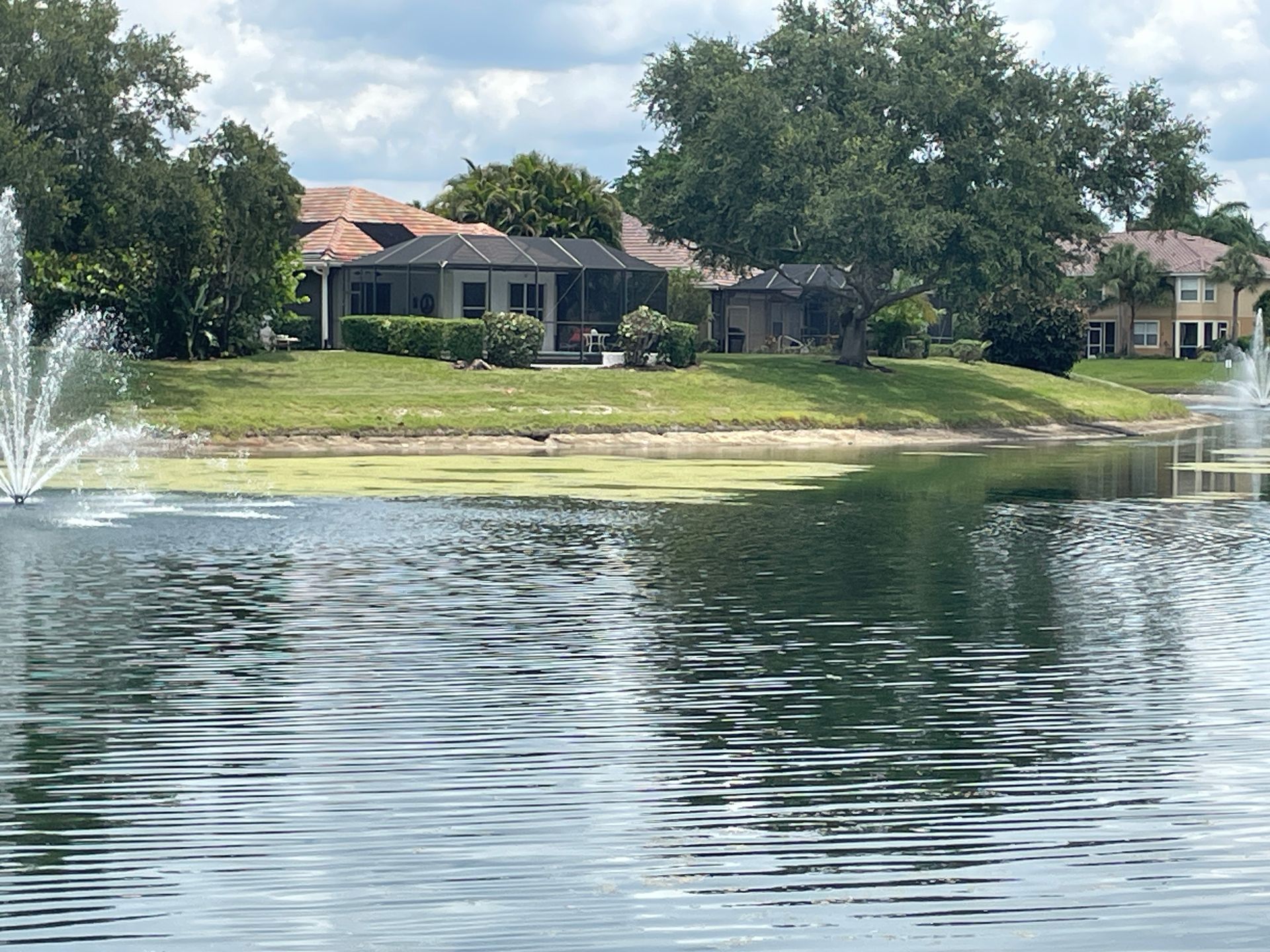 Residential homes beside a calm lake with fountains and green trees under a cloudy sky