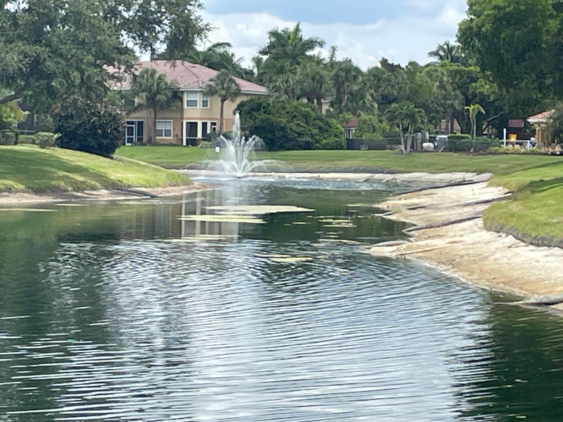 Pond with a fountain beside manicured lawns, palm trees, and houses under a partly cloudy sky